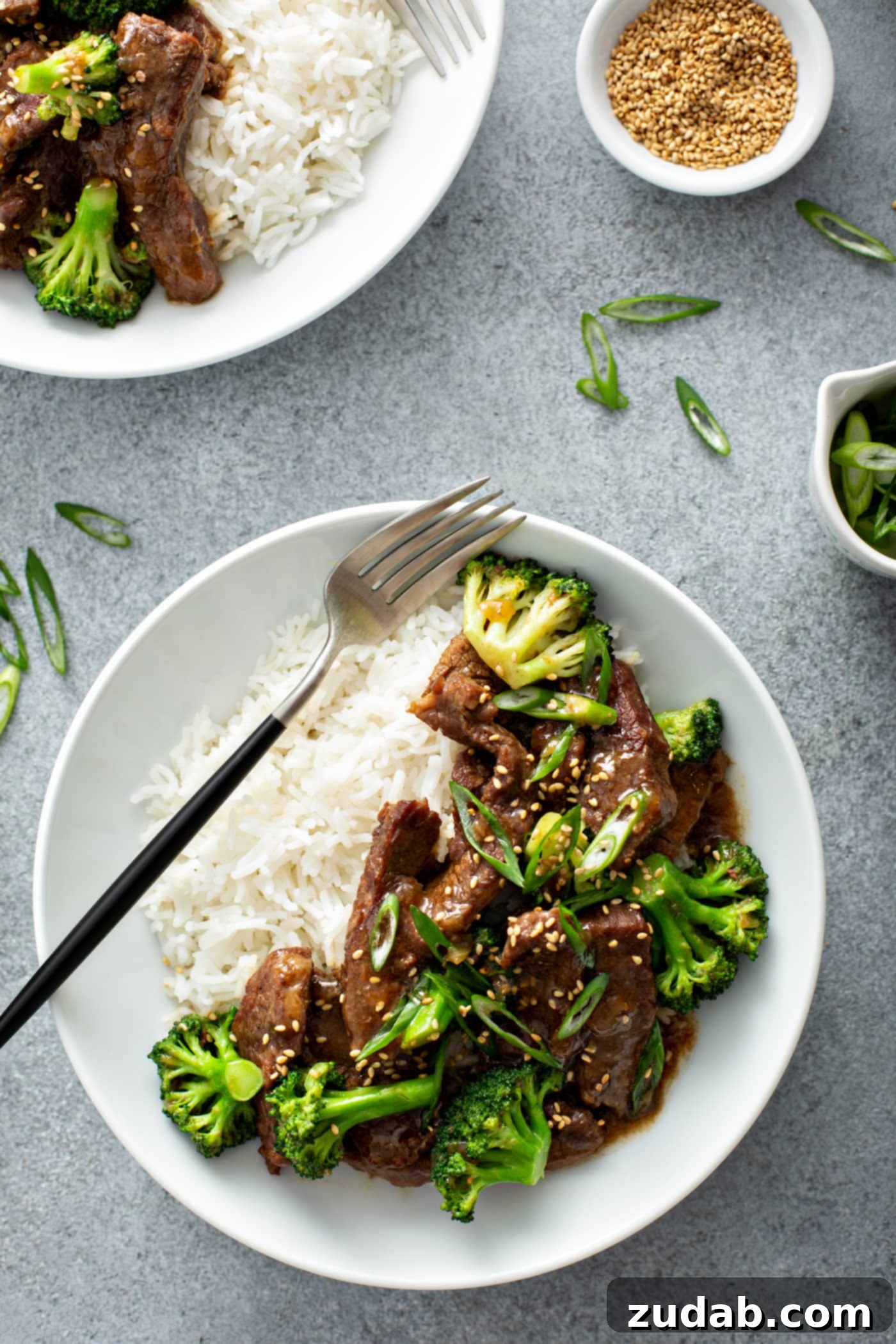 Instant Pot Beef and Broccoli A photo of a white bowl full of beef and broccoli and a side of white rice with a fork sitting on top of ther rice.