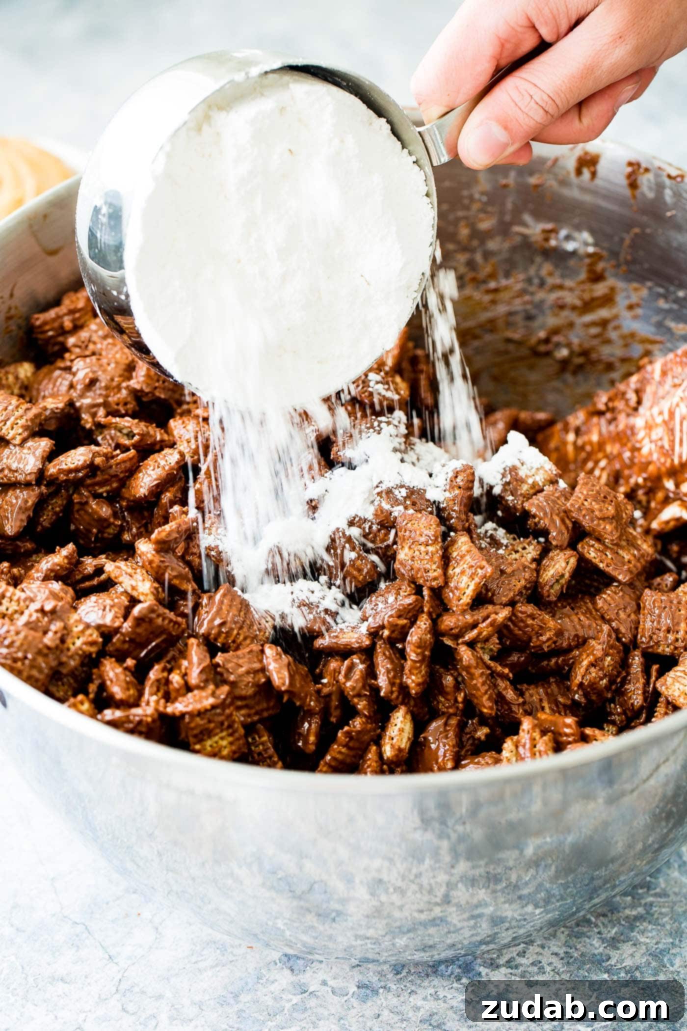 Chex Puppy Chow Muddy Buddies Mix A photo of powdered sugar being add to Chex cereal that has been coated with melted chocolate and peanut butter.