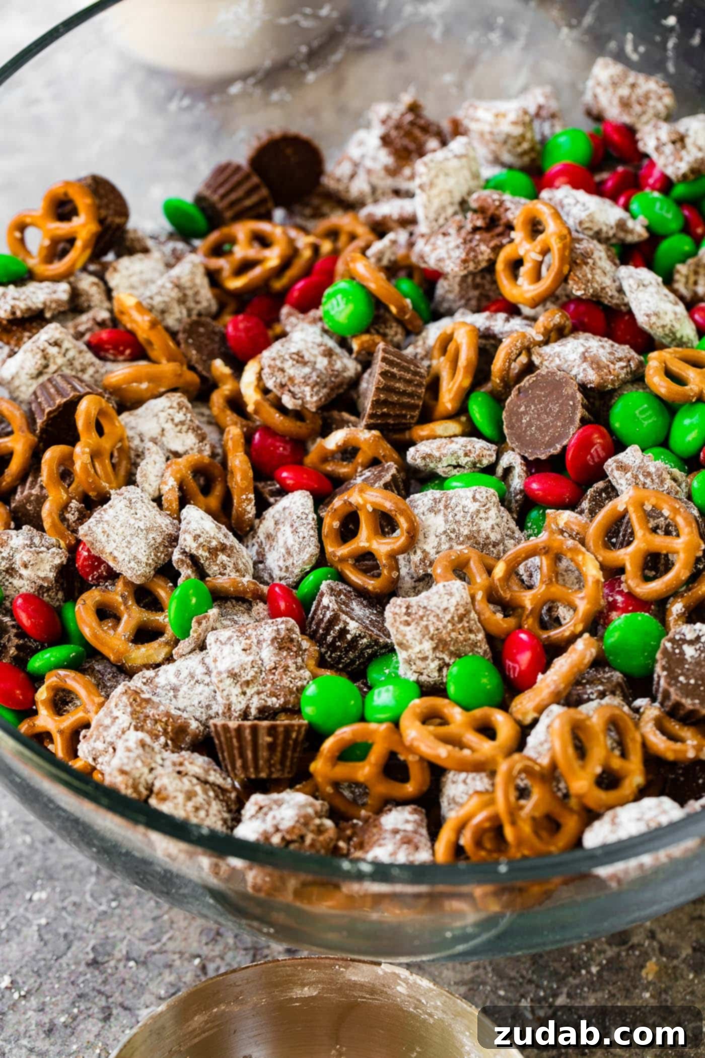 Reindeer Muddy Buddies Christmas Snack Mix A large glass bowl filled with a festive Christmas snack mix featuring pretzels, colorful M&Ms, rich peanut butter cups, and classic Chex muddy buddies, perfect for holiday gatherings.