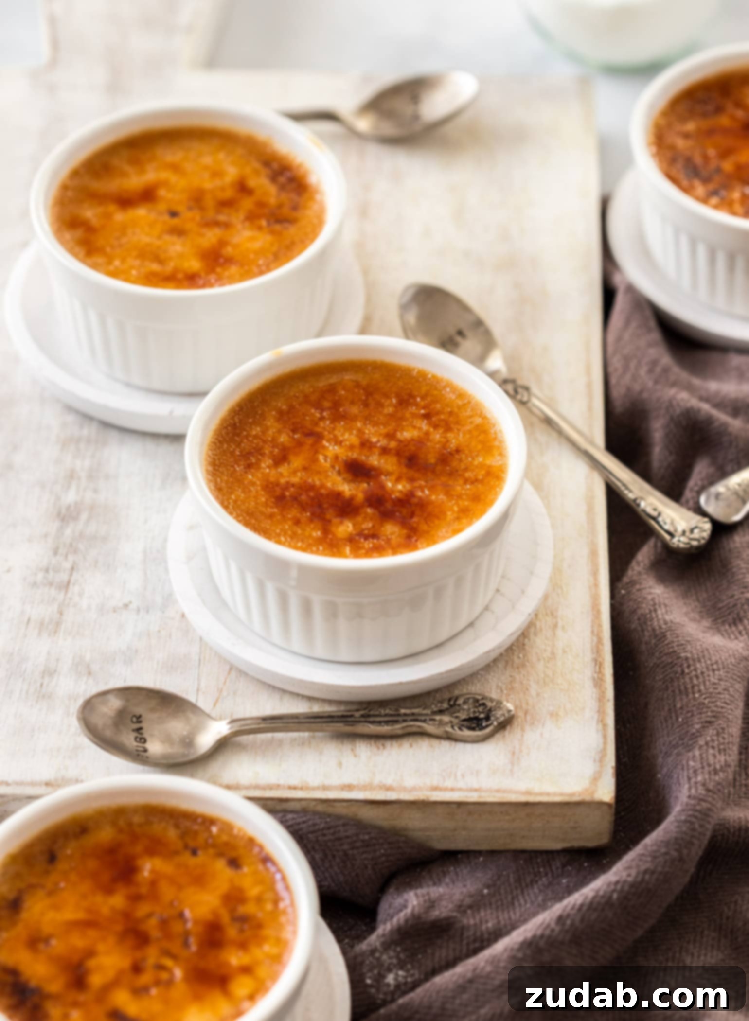 Four pristine white ramekins, each filled with expertly prepared crème brûlée, spoons resting beside them, elegantly arranged on a bright white serving board.