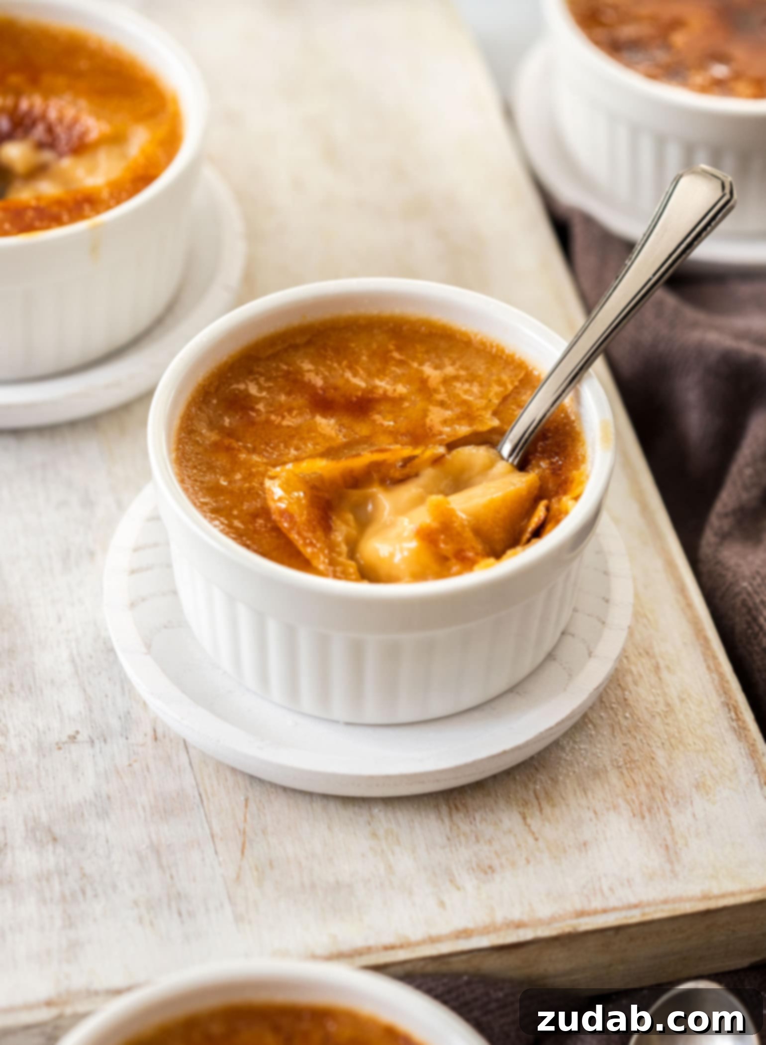 Four small, elegant white ramekins, each containing a finished salted caramel crème brûlée, with spoons arranged around them on a pristine white surface, ready for serving.