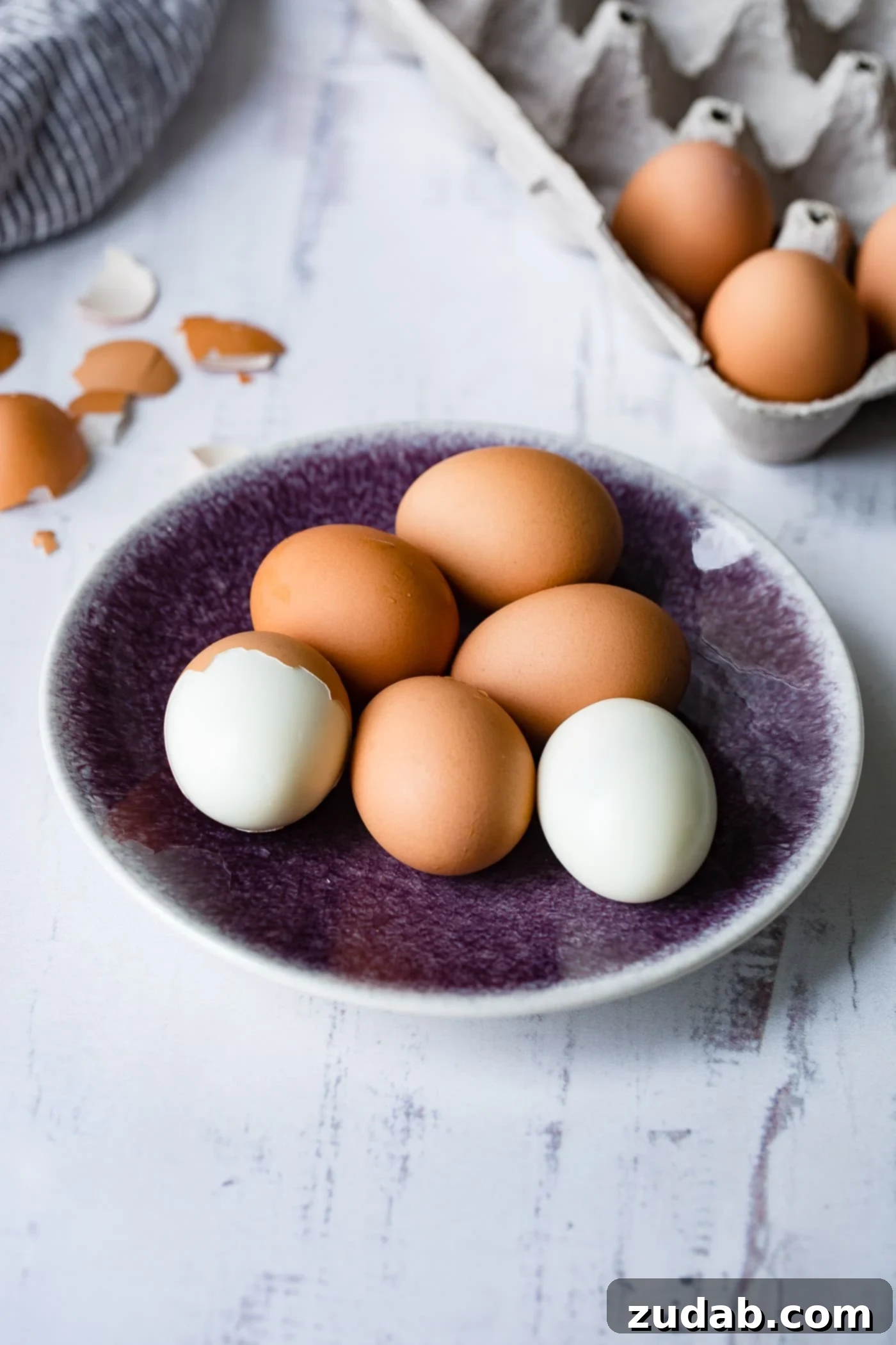 Instant Pot Hard Boiled Eggs A plate with 6 brown hard boiled eggs. One has been peeled and one is partially peeled. There are brown egg shell and a carton of eggs in the background.
