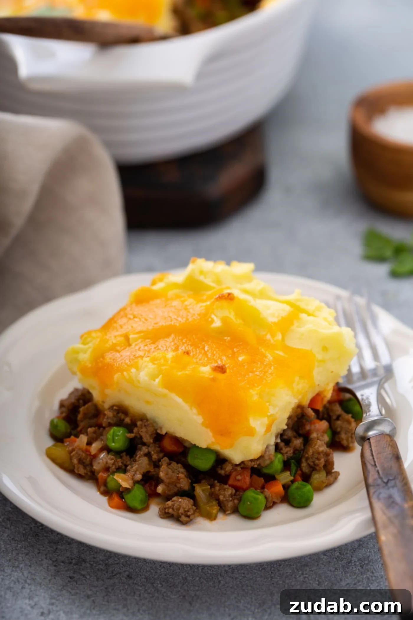 Classic Shepherd's Pie A serving of shepherd's pie on a white plate with a fork beside it. There is a napkin and a casserole dish in the background.