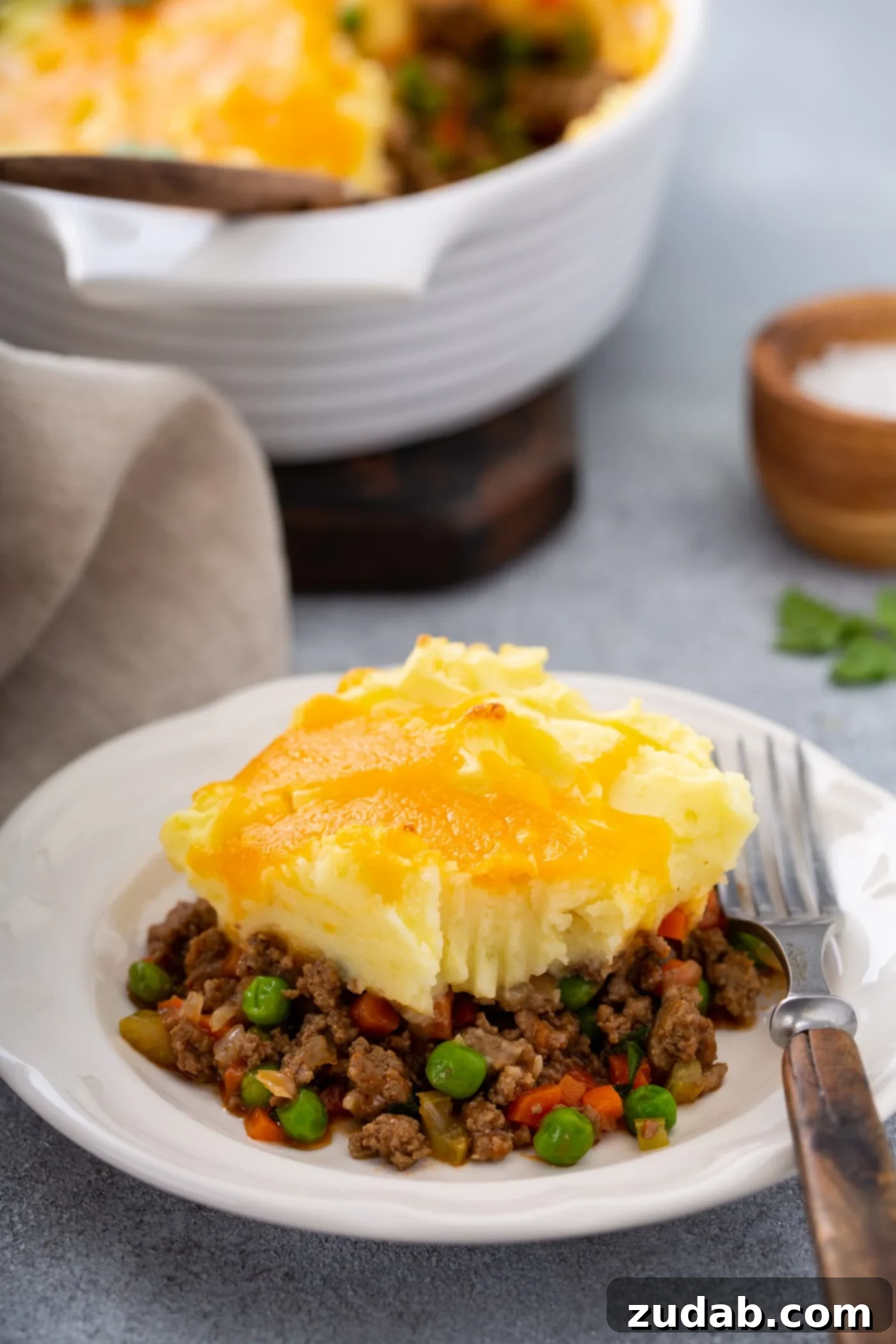 Classic Shepherd's Pie A serving of shepherd's pie with carrots, peas and ground beef for filling, topped with mashed potatoes and melted cheese. There is a fork resting on the plate and a napkin and a small container of salt next to it.
