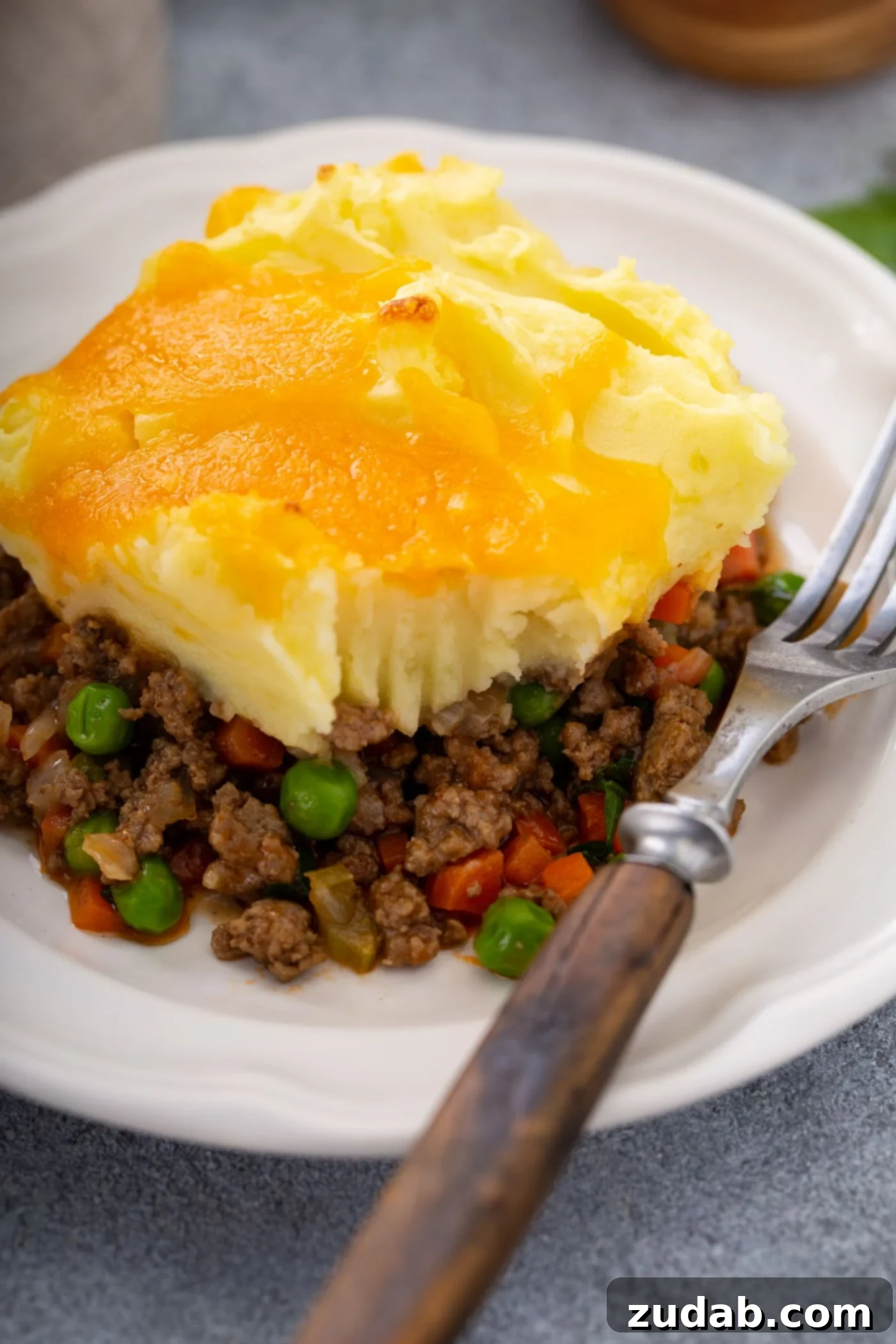 Classic Shepherd's Pie A serving of shepherd's pie on a white plate with a fork resting beside it. You can see the rich ground beef and vegetable filling, the mashed potatoes on top, and the yellow melted cheddar cheese topping it all.