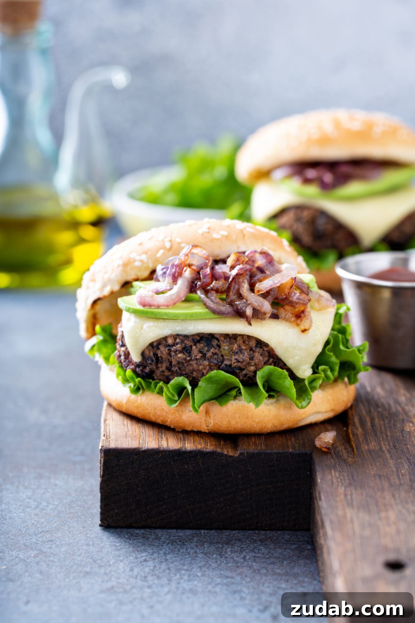 Your New Favorite Homemade Black Bean Burger 11 A table top showcases two perfectly prepared black bean burgers on toasted sesame seed buns. The burgers are generously topped with caramelized onions, creamy avocados, melted cheese, and fresh lettuce. In the soft background, a bowl of fresh lettuce and a container of cooking oil complete the inviting scene.