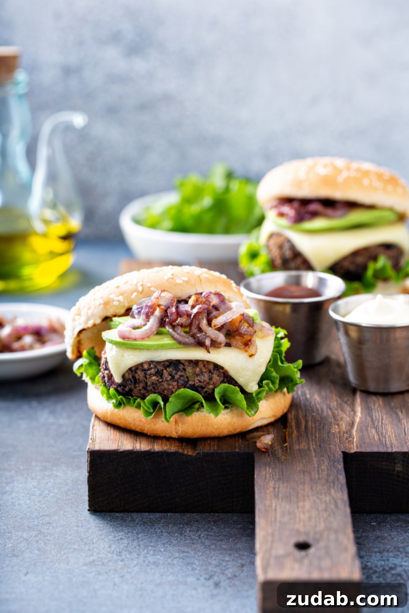 Your New Favorite Homemade Black Bean Burger 12 A perfectly assembled black bean burger on a sesame seed bun, richly topped with onions, creamy avocado, melted cheese, and crisp lettuce. In the background, another burger, a bowl of lettuce, a container of oil, a dish of sliced onions, and a container of mayo suggest a delightful meal spread.