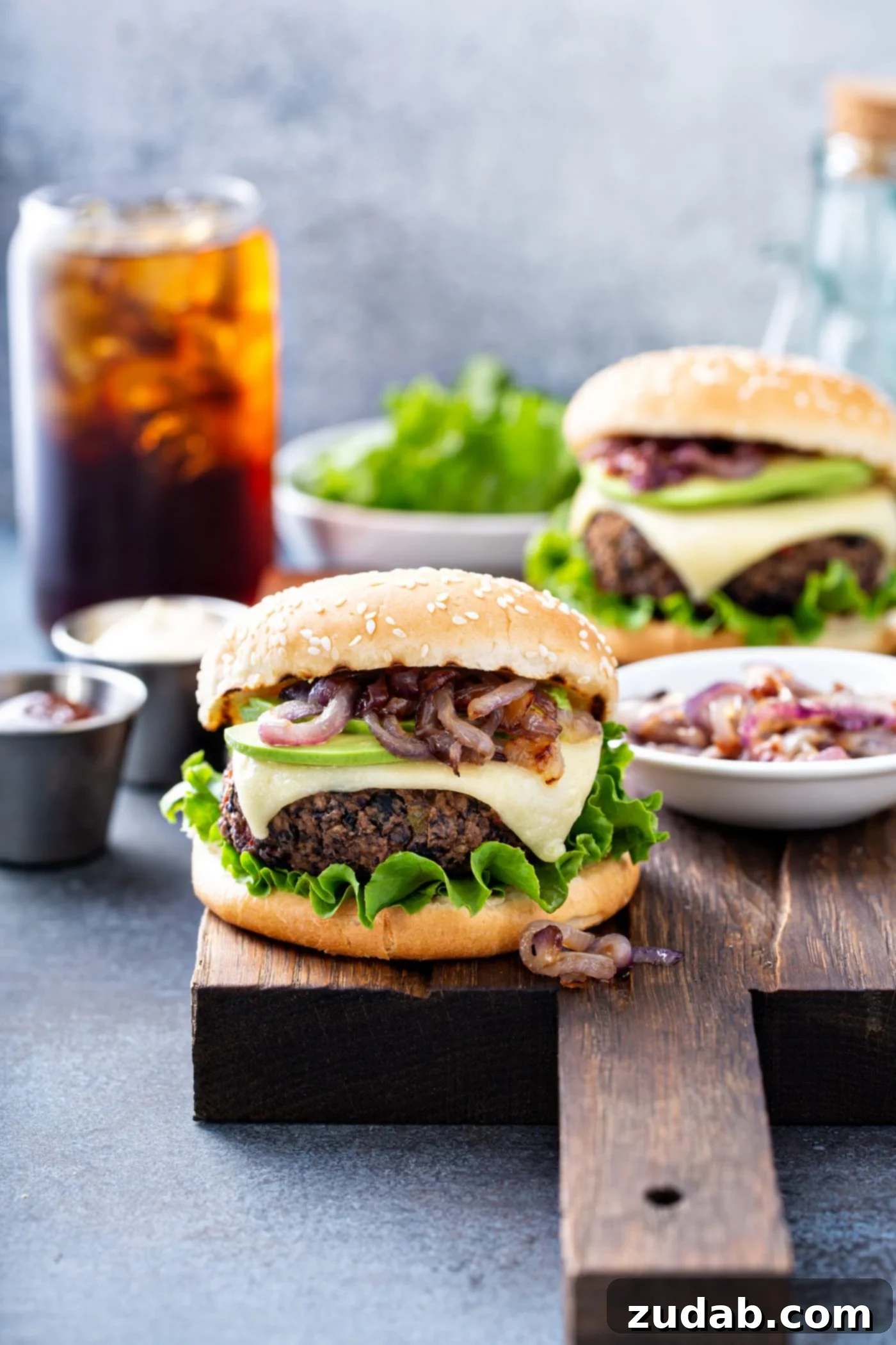 Your New Favorite Homemade Black Bean Burger 3 A black bean burger on a sesame seed bun topped with onions, avocado, cheese and lettuce. There is another burger, a bowl of lettuce, a container of onions, a container of mayo and a glass of iced soda in the background, hinting at a full meal spread.