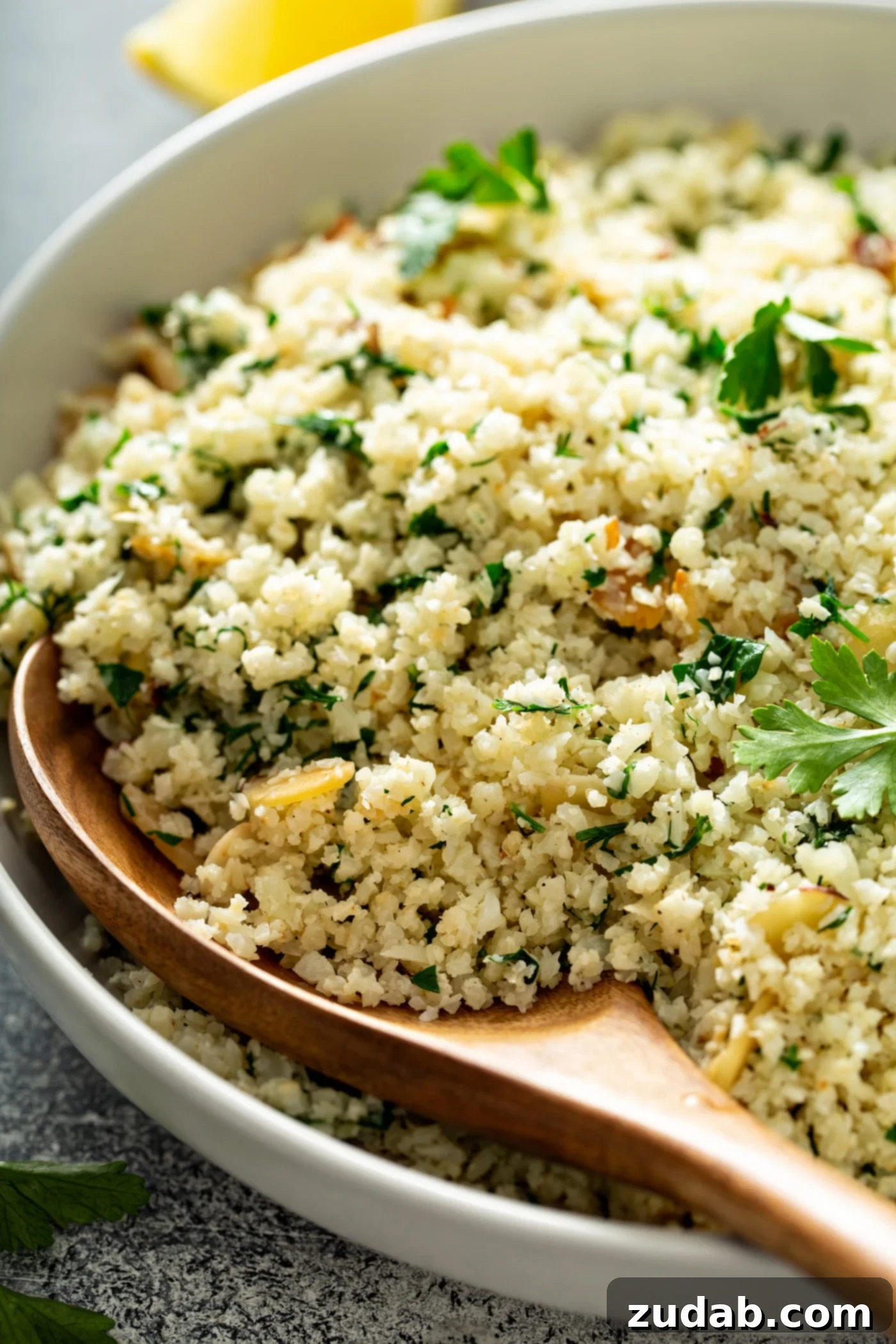 Garlic Herb Cauliflower Rice: A Healthy and Flavorful Side Dish A serving bowl filled with garlic herb cauliflower rice. You can see the green herbs in the rice and there is a wooden spoon filled with cauliflower rice resting in the bowl. Fresh parsley leaves are scattered on top.