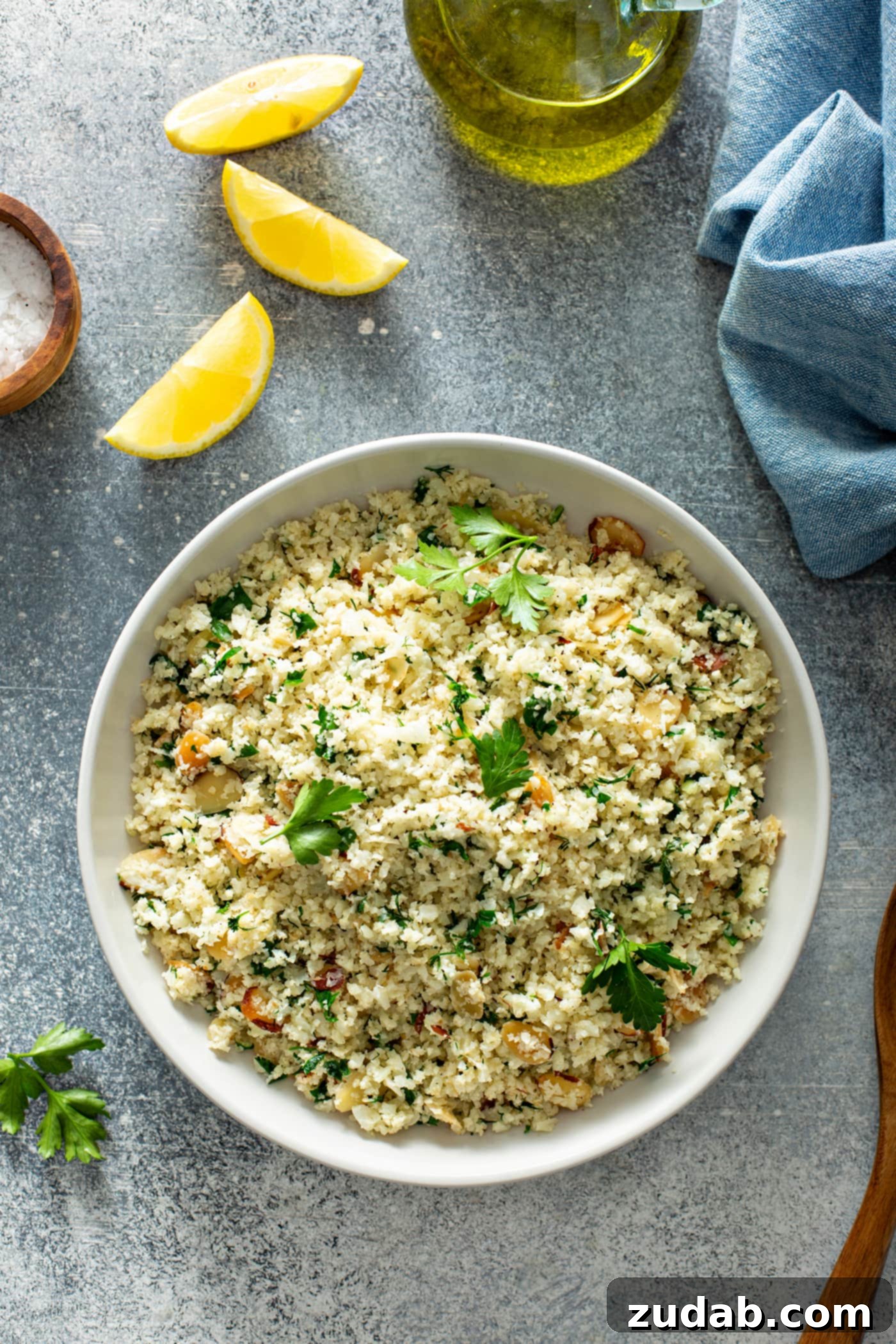 Flavorful Garlic Herb Cauliflower Rice with Fresh Garnish A white serving bowl filled with garlic herb cauliflower rice. Green herbs and sliced almonds are visible throughout the rice. In the soft-focus background, there's a container of salt, three bright lemon wedges, a glass container of oil, and a blue linen napkin, suggesting a fresh and prepared culinary setting.