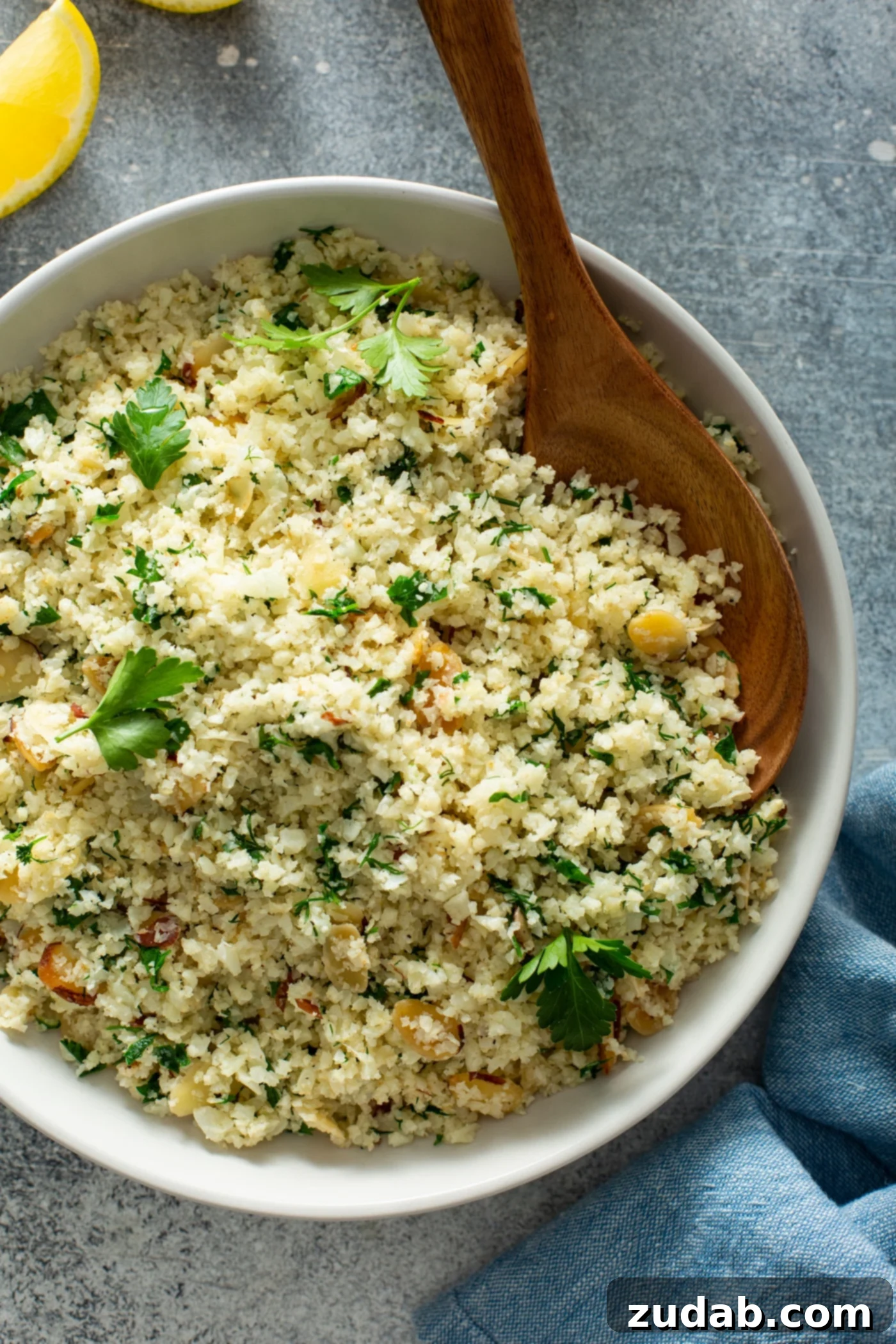 Healthy Garlic Herb Cauliflower Rice Ready to Serve A pristine white serving bowl filled with garlic herb cauliflower rice. The rice is visibly speckled with green herbs and golden sliced almonds, and garnished with fresh parsley leaves scattered artistically on top, making it an inviting and healthy dish.