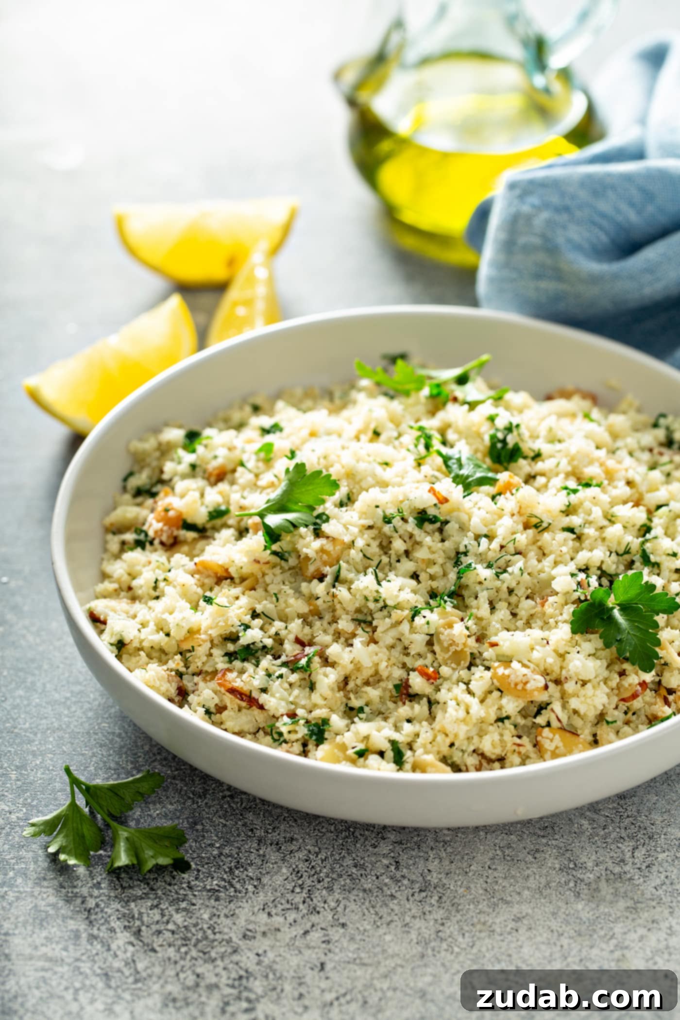 Delicious Garlic Herb Cauliflower Rice for Healthy Eating A white serving bowl containing garlic herb cauliflower rice. There are green herbs, garlic and sliced almonds in the rice. Parsley leaves are scattered on top of the rice. There are four lemon wedges next to the bowl of rice, enhancing the visual appeal.