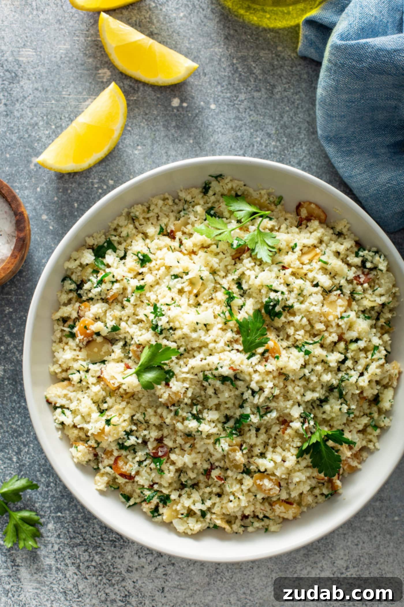 Serving Garlic Herb Cauliflower Rice with Lemon A white serving bowl of garlic herb cauliflower rice, meticulously topped with fresh parsley leaves. In the background, there are bright lemon wedges, a small container of sea salt, and a soft blue linen napkin, adding to the serene culinary presentation.