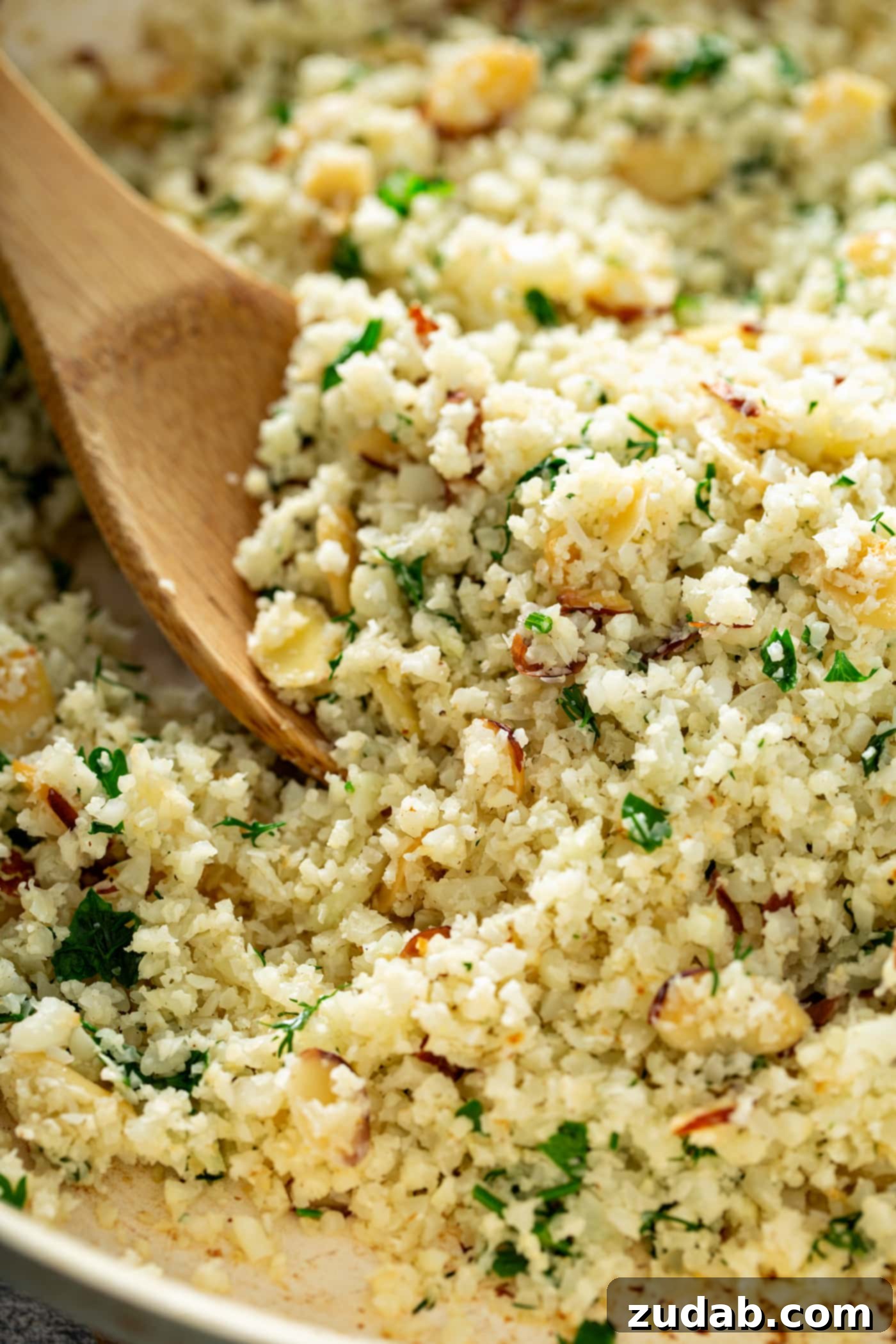 Close-up of Garlic Herb Cauliflower Rice with Serving Spoon A close-up shot of a bowl of garlic herb cauliflower rice, featuring a wooden serving spoon nestled within. You can clearly see the vibrant green herbs and scattered sliced almonds throughout the fluffy rice, highlighting its texture and freshness.