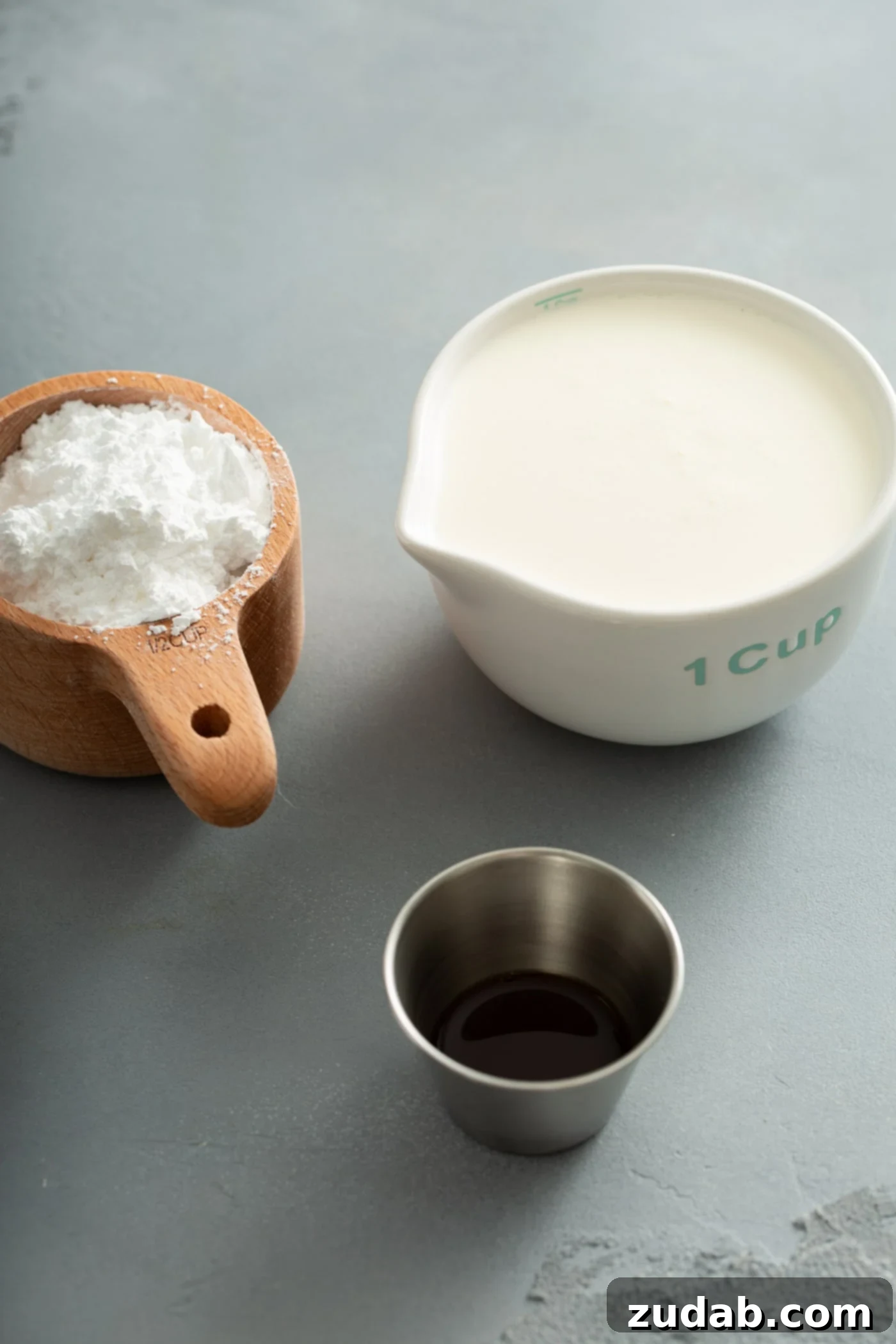 Simply Delicious Homemade Whipped Cream 3 A measuring cup of heavy cream, a measuring cup containing powdered sugar, and a small silver container of vanilla extract are neatly arranged on a white surface, showcasing the minimal ingredients needed for homemade whipped cream.