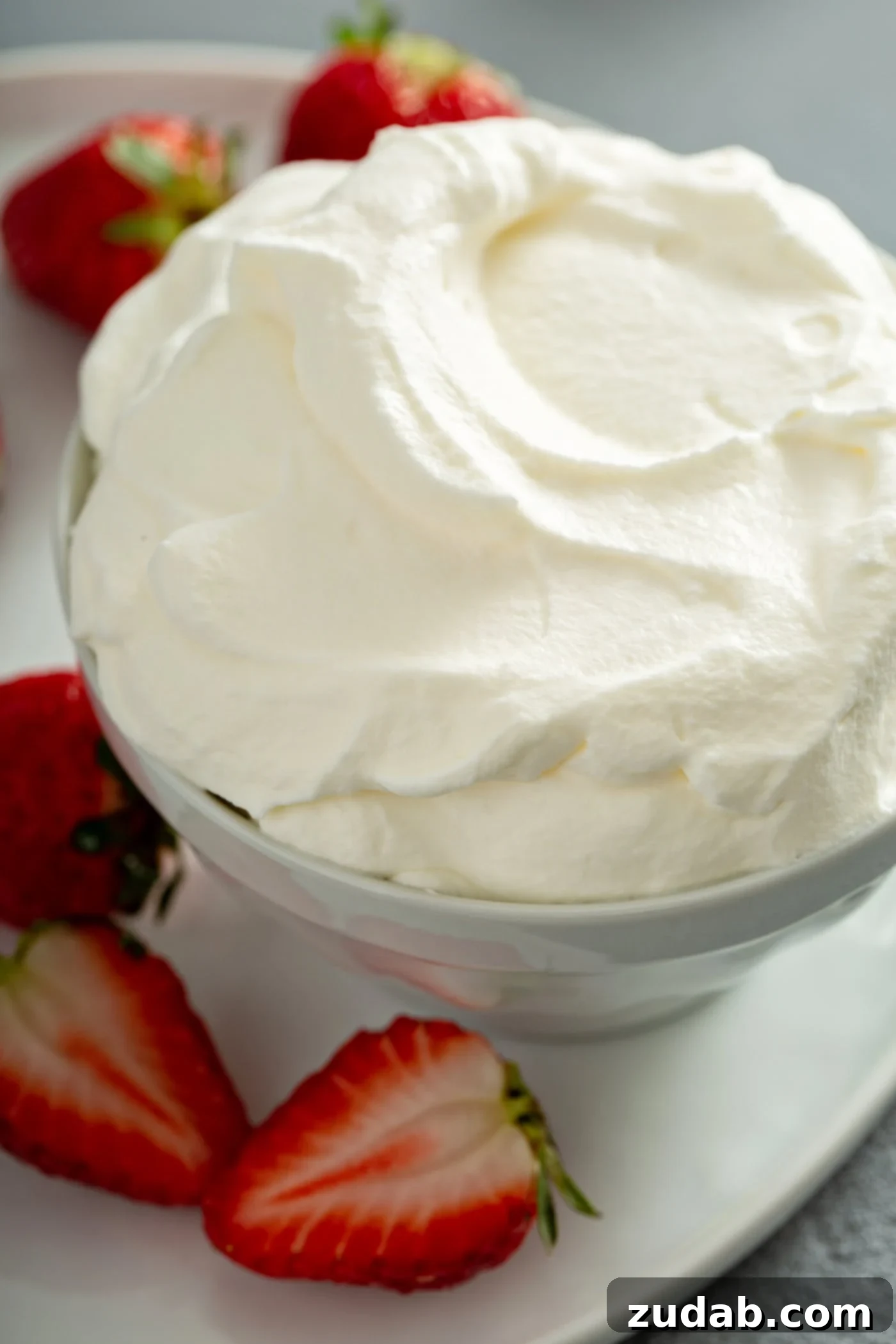 Simply Delicious Homemade Whipped Cream 5 A close-up of a bowl of fluffy homemade whipped cream, elegantly swirled and garnished with fresh, ripe strawberries, highlighting its appeal as a dessert topping.