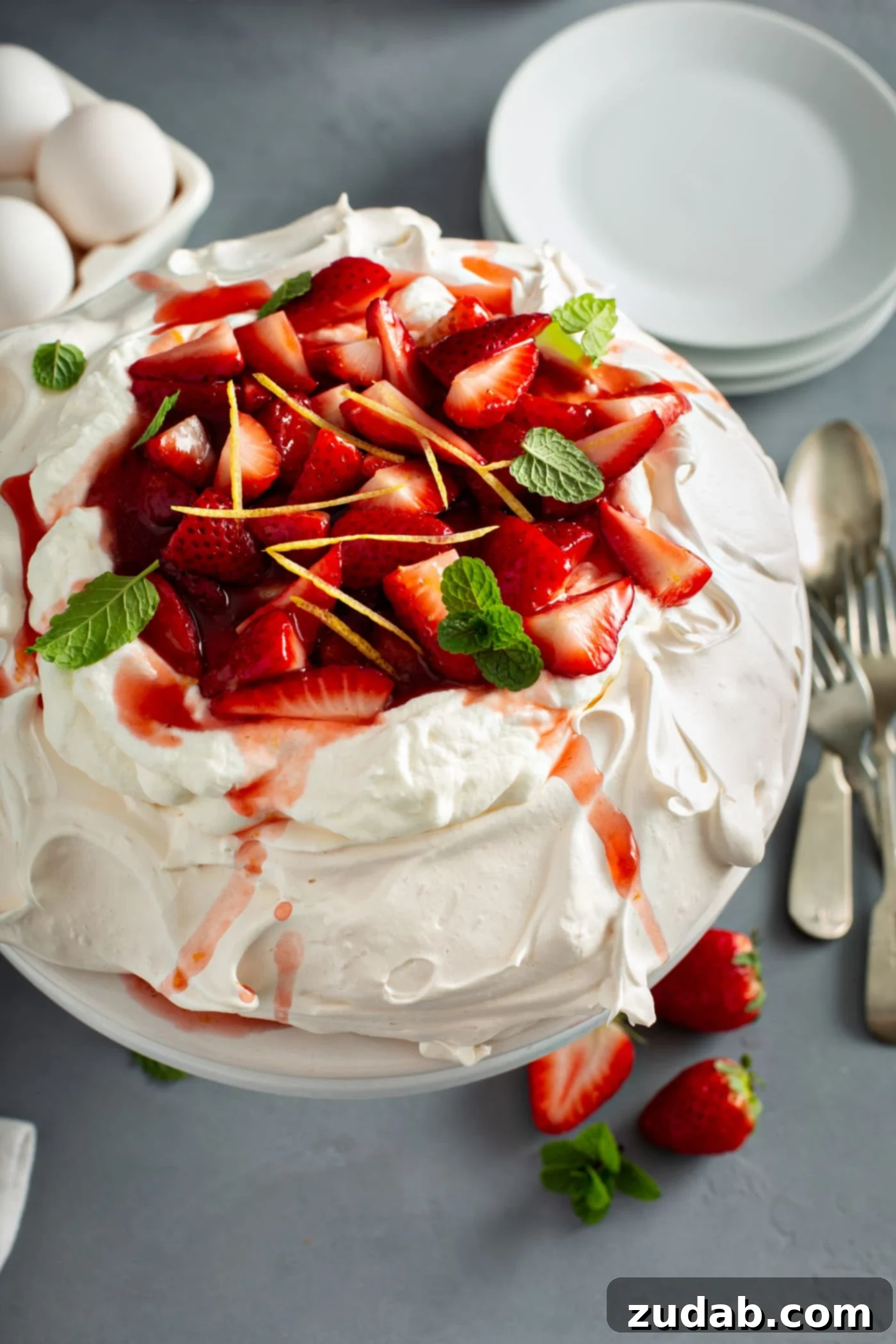 Pavlova topped with sliced strawberries, slivered lemon rind, and mint leaves. A fork and spoon and some sliced strawberries lay beside the pavlova. And there are white dessert plates in the background.