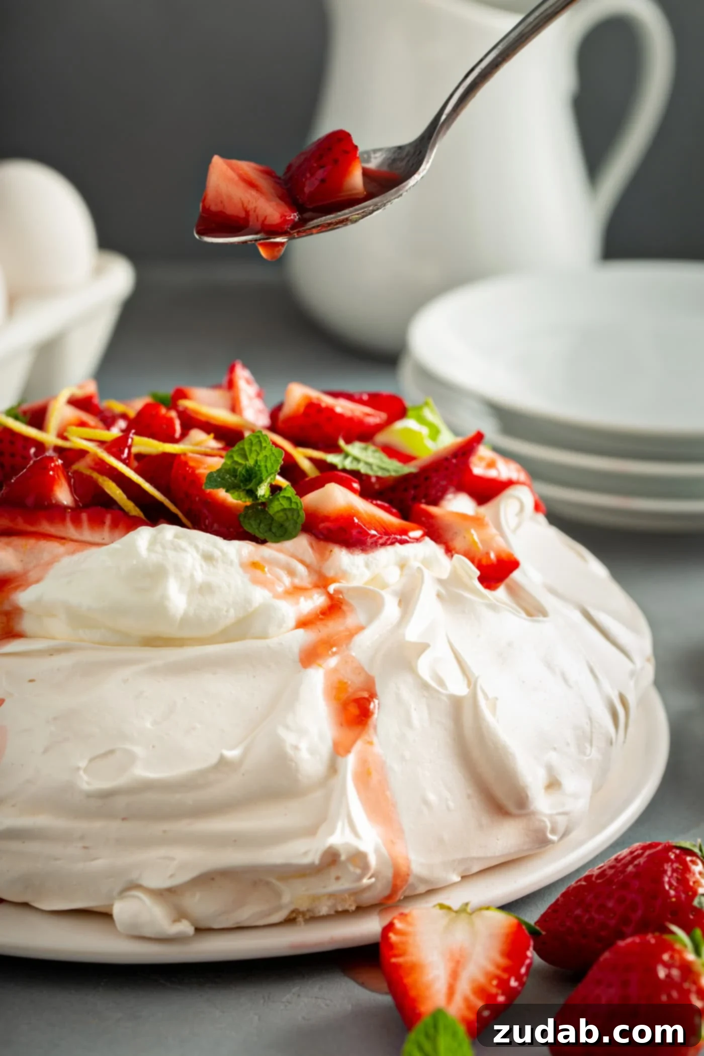 Pavlova on a serving plate. It is topped with whipped cream, sliced strawberries, slivers of lemon rind, and a few mint leaves. A spoon with strawberries and juice drizzling down is above the pavlova. There are white dessert plates, eggs, 