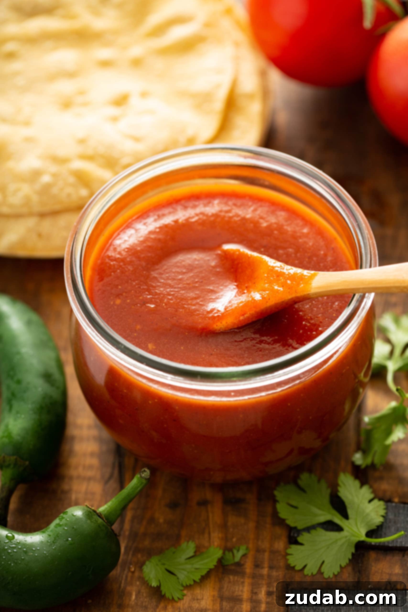 Homemade Red Enchilada Sauce A glass container with a wooden spoon. The container is filled with rich red enchilada sauce, ready for use. Tortillas, green chilies, and fresh cilantro leaves are arranged around the container, hinting at a delicious meal.