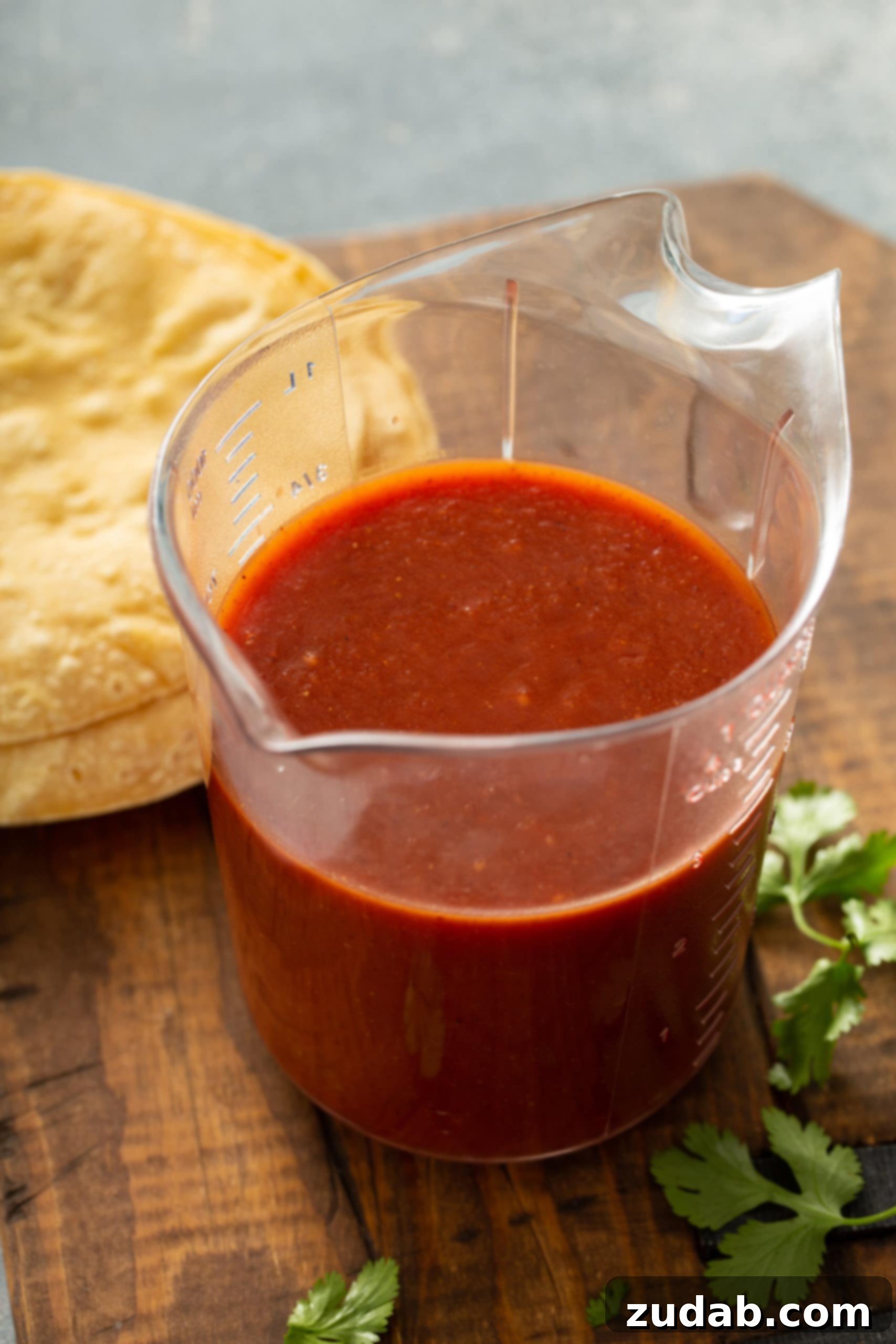 Homemade Red Enchilada Sauce A glass measuring cup filled with vibrant red enchilada sauce, ready to be poured over delicious enchiladas. In the background, a neat stack of corn tortillas and a few fresh cilantro leaves complete the appetizing scene.