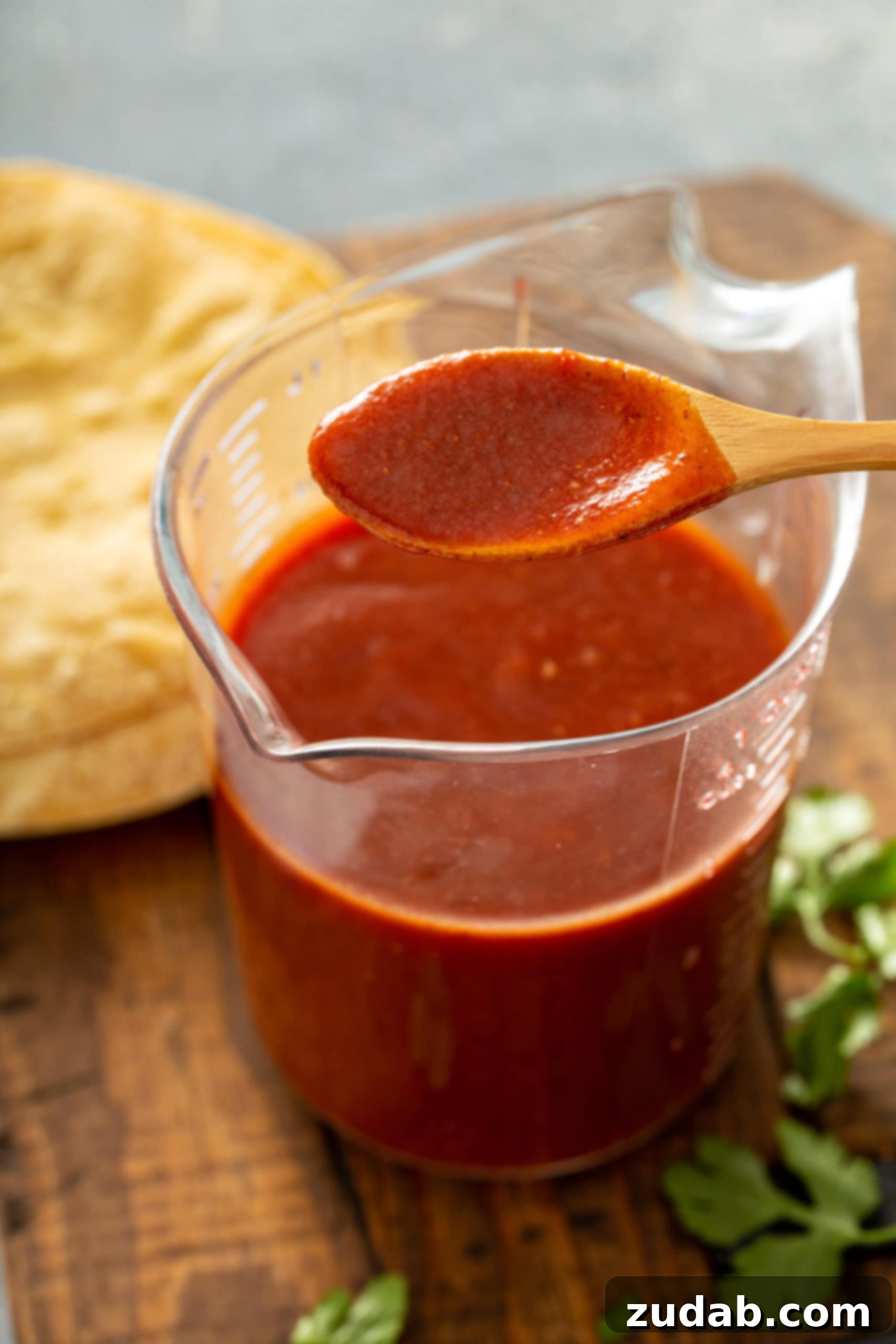 Homemade Red Enchilada Sauce A glass measuring cup brimming with rich red enchilada sauce, with a wooden spoon dipped into its velvety texture. In the soft-focus background, a stack of tortillas and fresh cilantro leaves hint at the delicious meal to come.