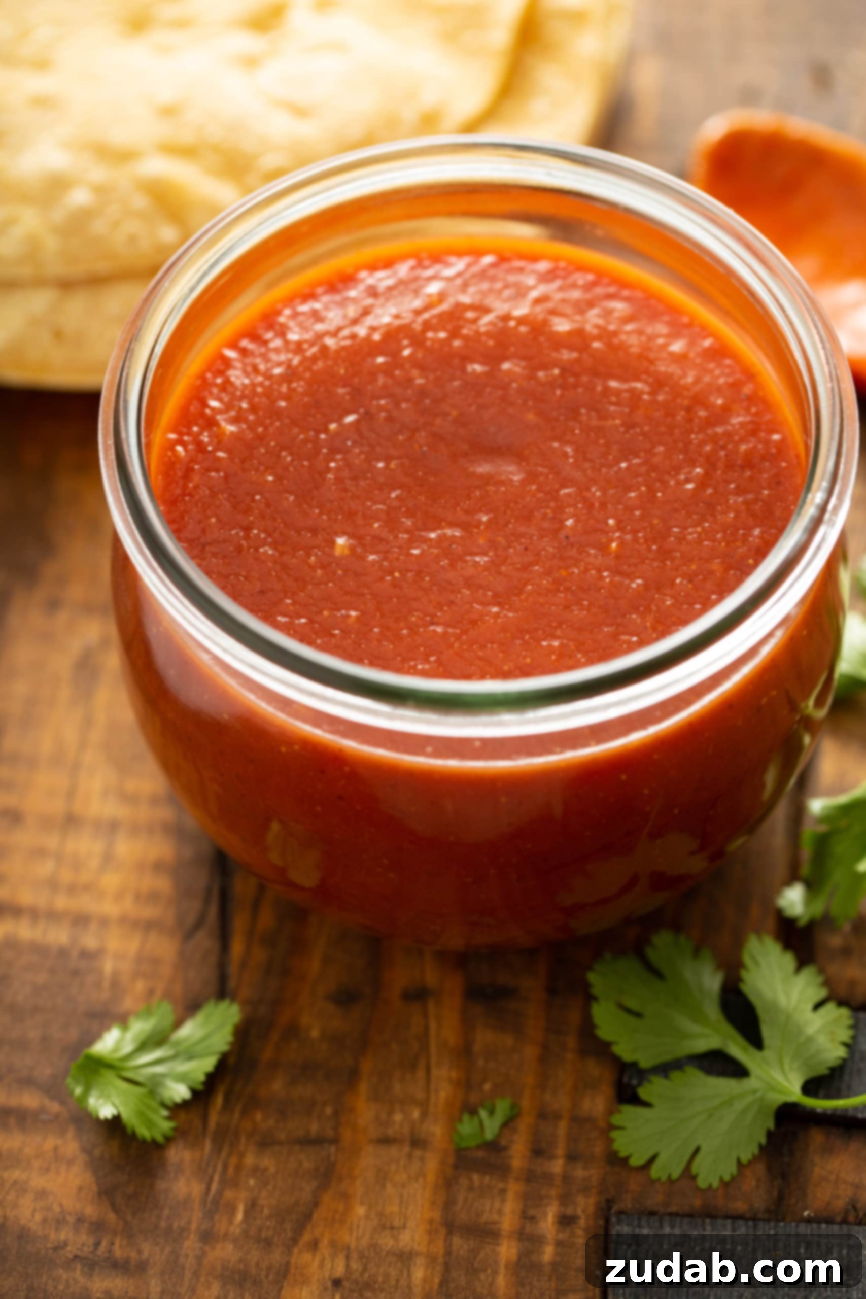 Classic Homemade Red Enchilada Sauce 2 A stylish glass jar generously filled with rich, homemade red enchilada sauce, invitingly placed. Surrounding it are fresh tortillas, vibrant cilantro, and ripe tomatoes, signaling the delicious, authentic Mexican flavors ready to be created.