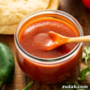A glass container with a wooden spoon. The container is filled with red enchilada sauce. There are tortillas, green chilies, and cilantro leaves surrounding the container of red sauce.