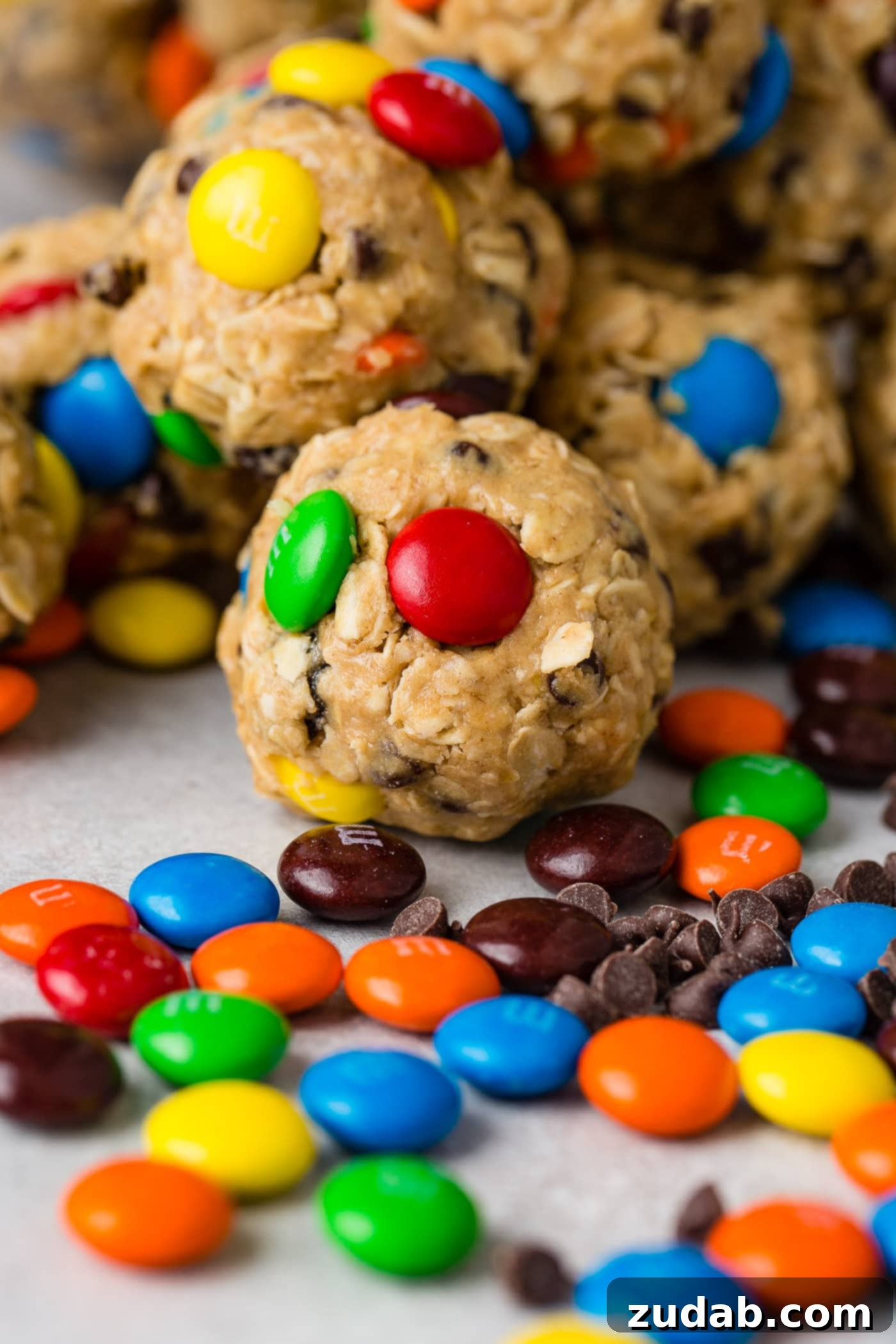 Loaded Peanut Butter Protein Bites 4 Seven vibrant protein energy balls, brimming with oats, raisins, mini M&Ms, and mini chocolate chips. Mini M&Ms and raisins are artfully scattered on the table in front of the protein balls, adding to their visual appeal.