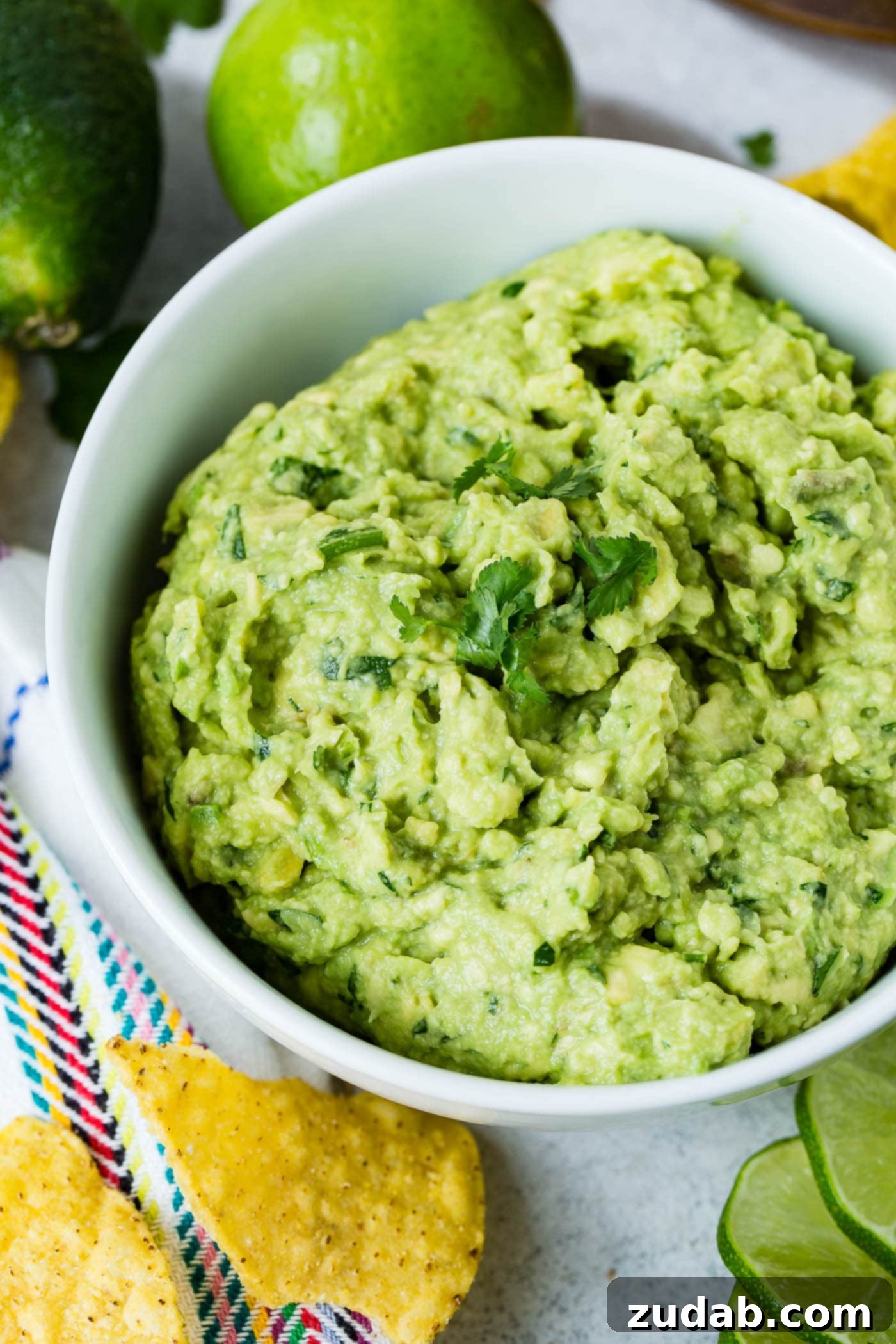 The Ultimate Homemade Authentic Guacamole Recipe 2 A white bowl of fresh guacamole. You can see the cilantro in the guacamole and a few cilantro leaves are on top. There are a few chips on the table next to the bowl.