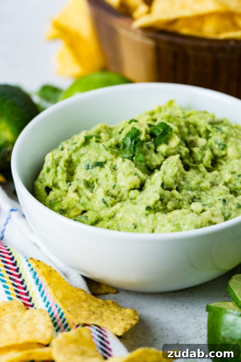 The Ultimate Homemade Authentic Guacamole Recipe 4 A white bowl of fresh guacamole with cilantro leaves on top. There are two whole limes and a wooden bowl of tortilla chips in the background. There are some more chips and slices of lime in front of the bowl.