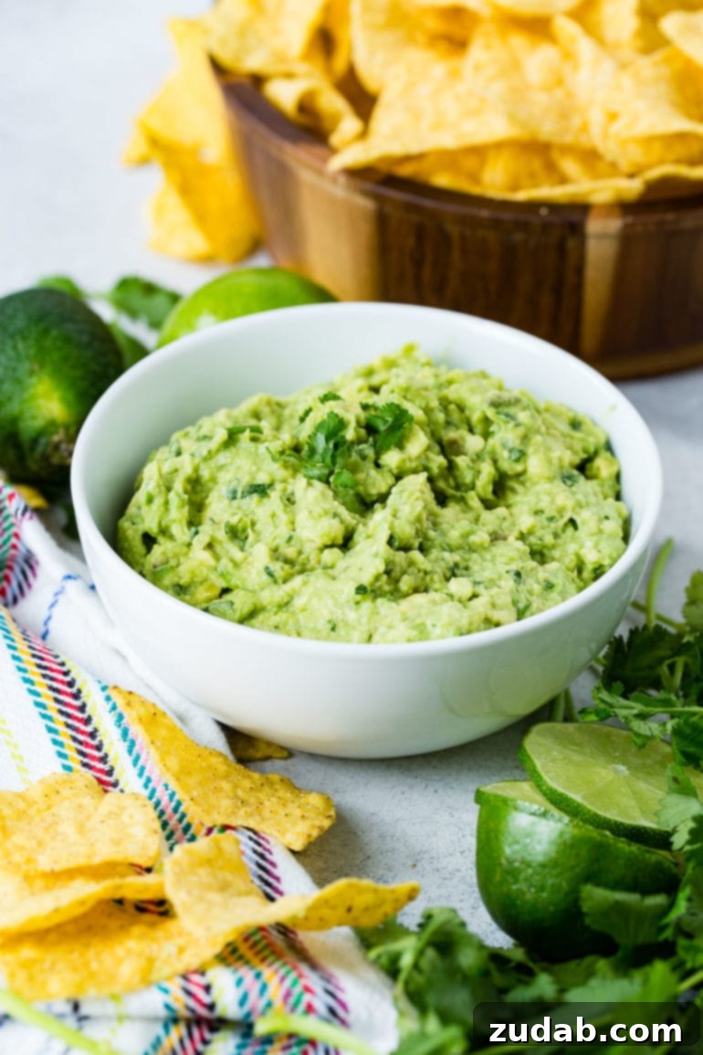 The Ultimate Homemade Authentic Guacamole Recipe 5 A bowl of fresh guacamole with a few cilantro leaves on top. There are tortilla chips scattered on the table in front of the bowl and a partially sliced lime next to the chips. There are two whole lime and a wooden bowl of tortilla chips in the background.