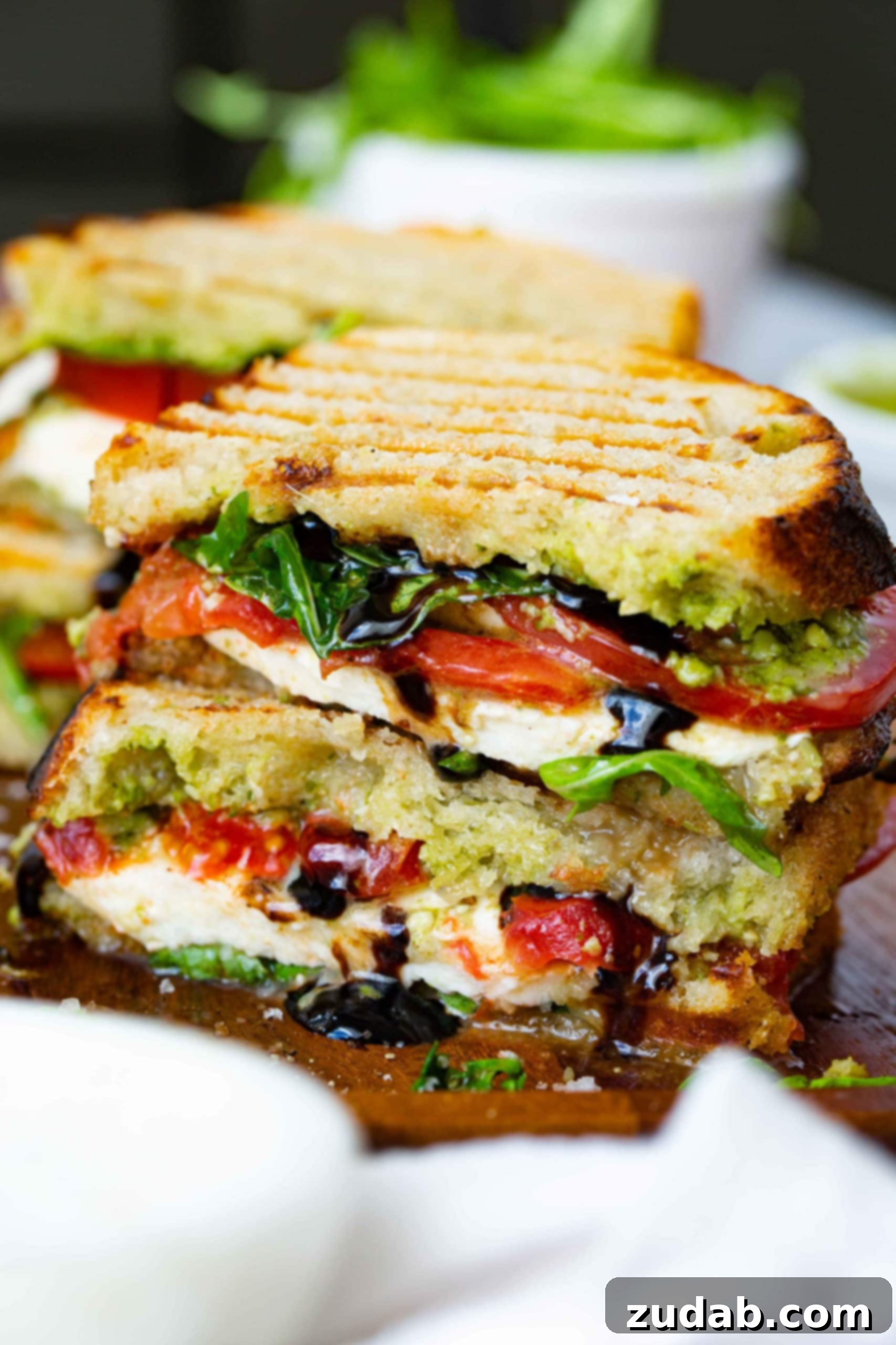 Tomato Mozzarella Basil Bliss 2 A perfectly grilled Balsamic Glazed Caprese Sandwich, cut in half and stacked. The golden-brown bread reveals layers of fresh mozzarella, ripe tomatoes, and arugula, with a rich balsamic glaze visibly drizzling out. Another complete sandwich is subtly blurred in the background, hinting at more deliciousness.