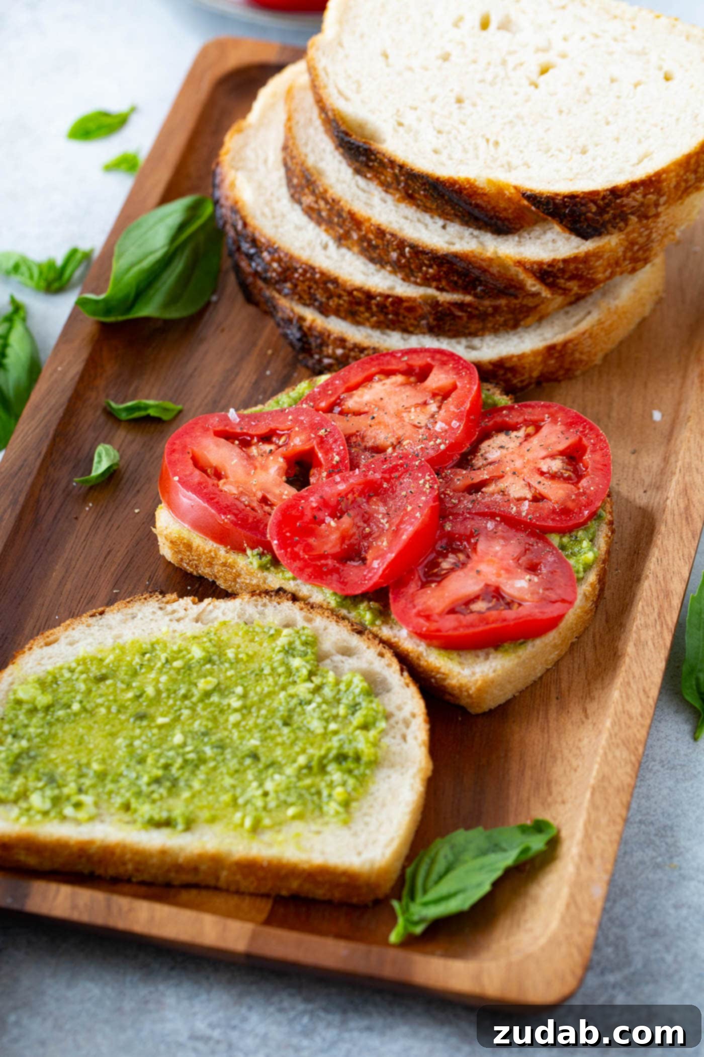 Tomato Mozzarella Basil Bliss 3 A beautifully arranged spread for making a Caprese Panini. Four slices of golden sourdough bread are stacked, with one slice separately topped with vibrant arugula and five slices of crimson tomatoes. Adjacent is another slice of bread generously spread with rich green basil pesto. Fresh basil leaves are artfully scattered across the rustic wooden tray, highlighting the premium, fresh ingredients.