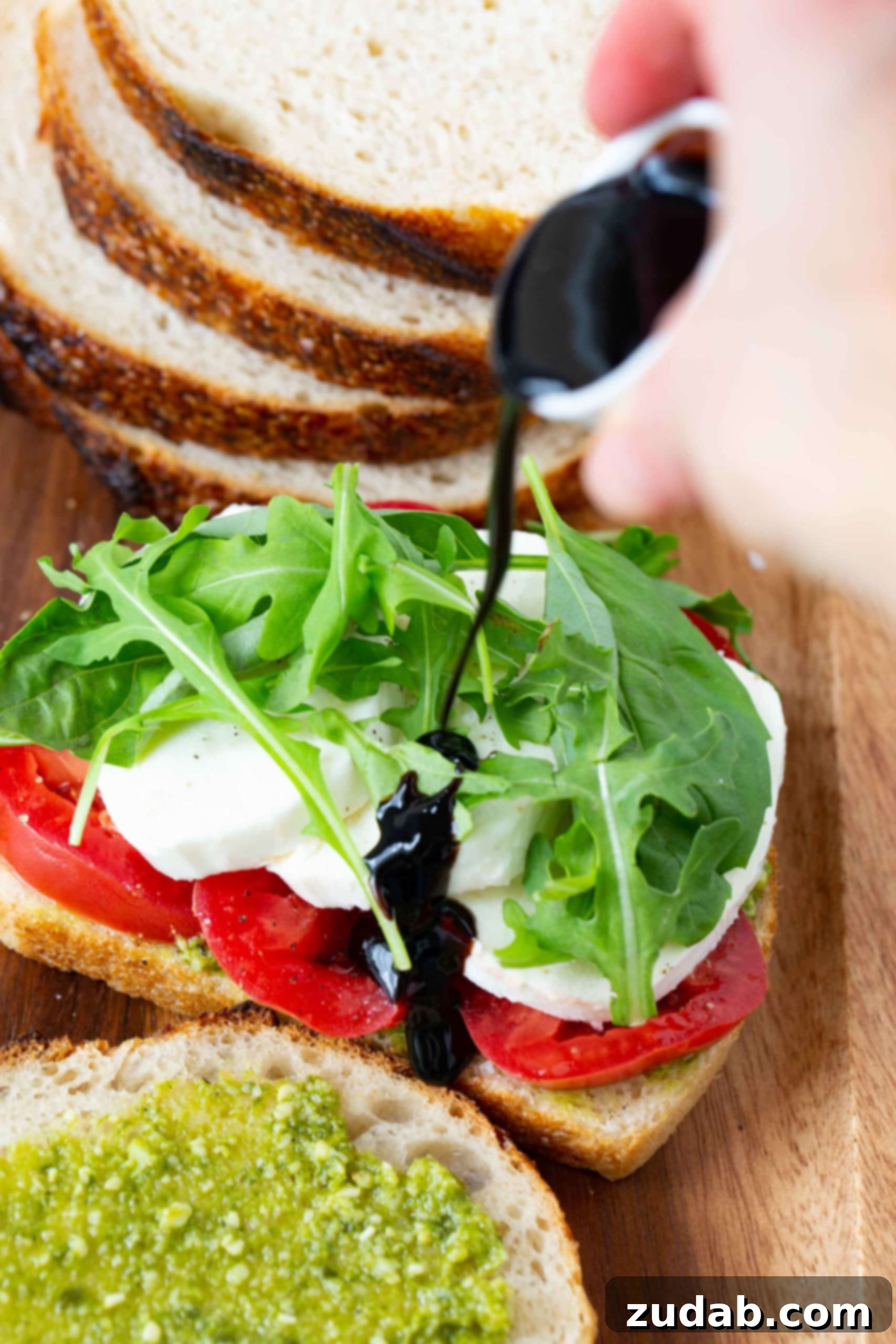 Tomato Mozzarella Basil Bliss 4 An open Caprese sandwich ready for grilling, showcasing layers of sliced tomatoes, fresh mozzarella cheese, and vibrant arugula. A luscious balsamic glaze is being artfully drizzled over the cheese, enhancing its appeal. In the foreground, a pesto-smeared slice of bread awaits, while stacked bread slices are visible in the background, signaling the sandwich-making process.