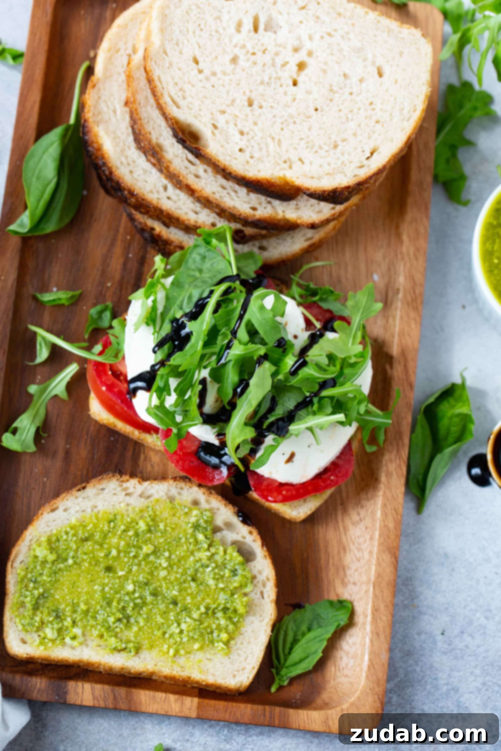 Tomato Mozzarella Basil Bliss 5 A beautifully composed Caprese Panini on a wooden tray, featuring a slice of sourdough bread generously topped with vibrant sliced tomatoes, creamy fresh mozzarella, and crisp arugula. Next to it, another slice of bread is spread with rich basil pesto. Scattered arugula and fresh basil leaves adorn the tray and table, emphasizing the freshness of the ingredients, ready for assembly.