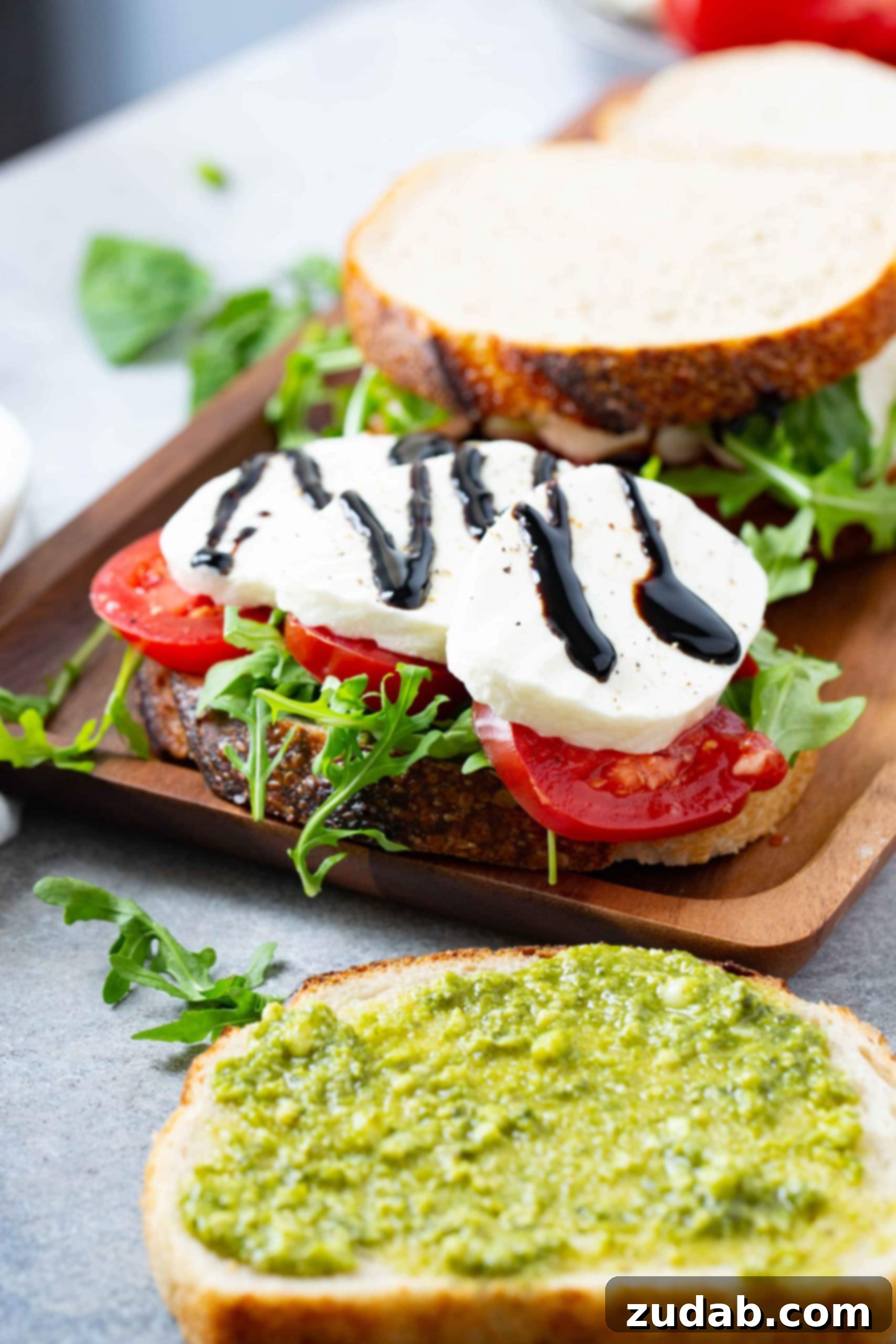 Tomato Mozzarella Basil Bliss 6 A close-up of a meticulously assembled Caprese sandwich on a wooden tray, featuring a slice of sourdough bread topped with vibrant sliced tomatoes, creamy fresh mozzarella, and crisp arugula. A glistening balsamic glaze is generously drizzled over the cheese. Additional slices of bread are stacked in the background, while loose arugula leaves and pesto-smeared bread are visible in the foreground, showcasing the fresh, inviting ingredients.