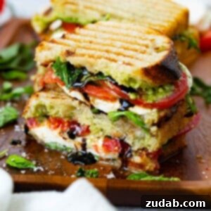 A grilled Caprese Panini, cut in half and stacked, showcasing grill marks and the delicious filling of arugula, tomatoes, pesto, and mozzarella cheese, with balsamic glaze oozing. A second sandwich and fresh ingredients are in the background.