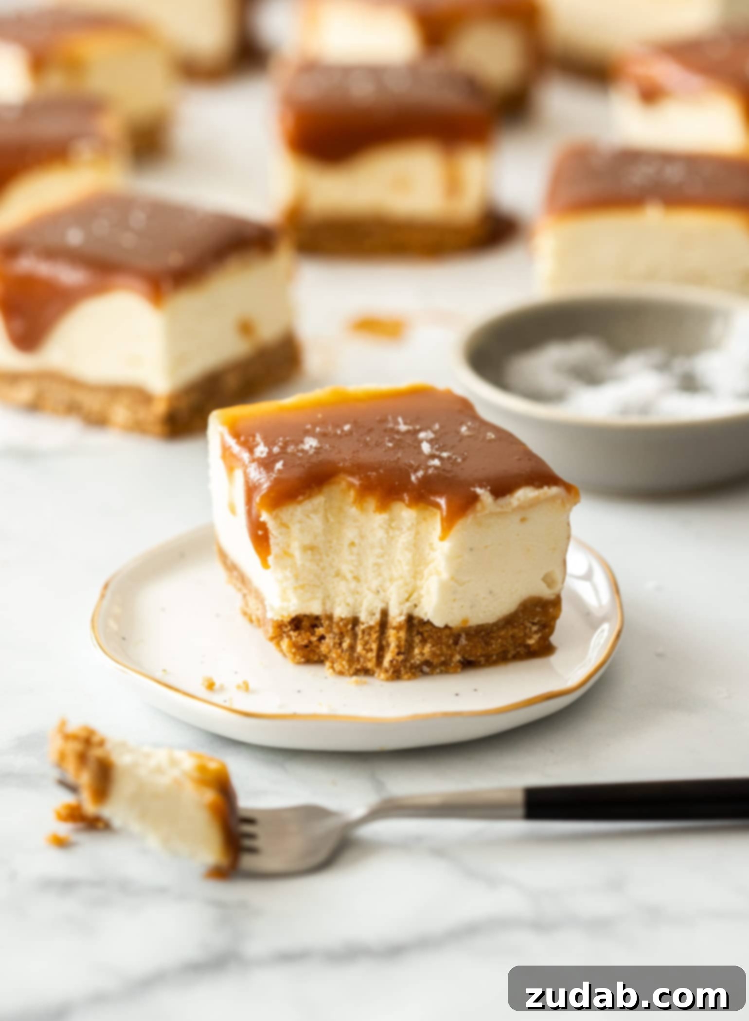 Effortless Caramel Bliss Bars 6 A single slice of No-Bake Caramel Cheesecake Bar on a small white plate with a fork, and other bars blurred in the background