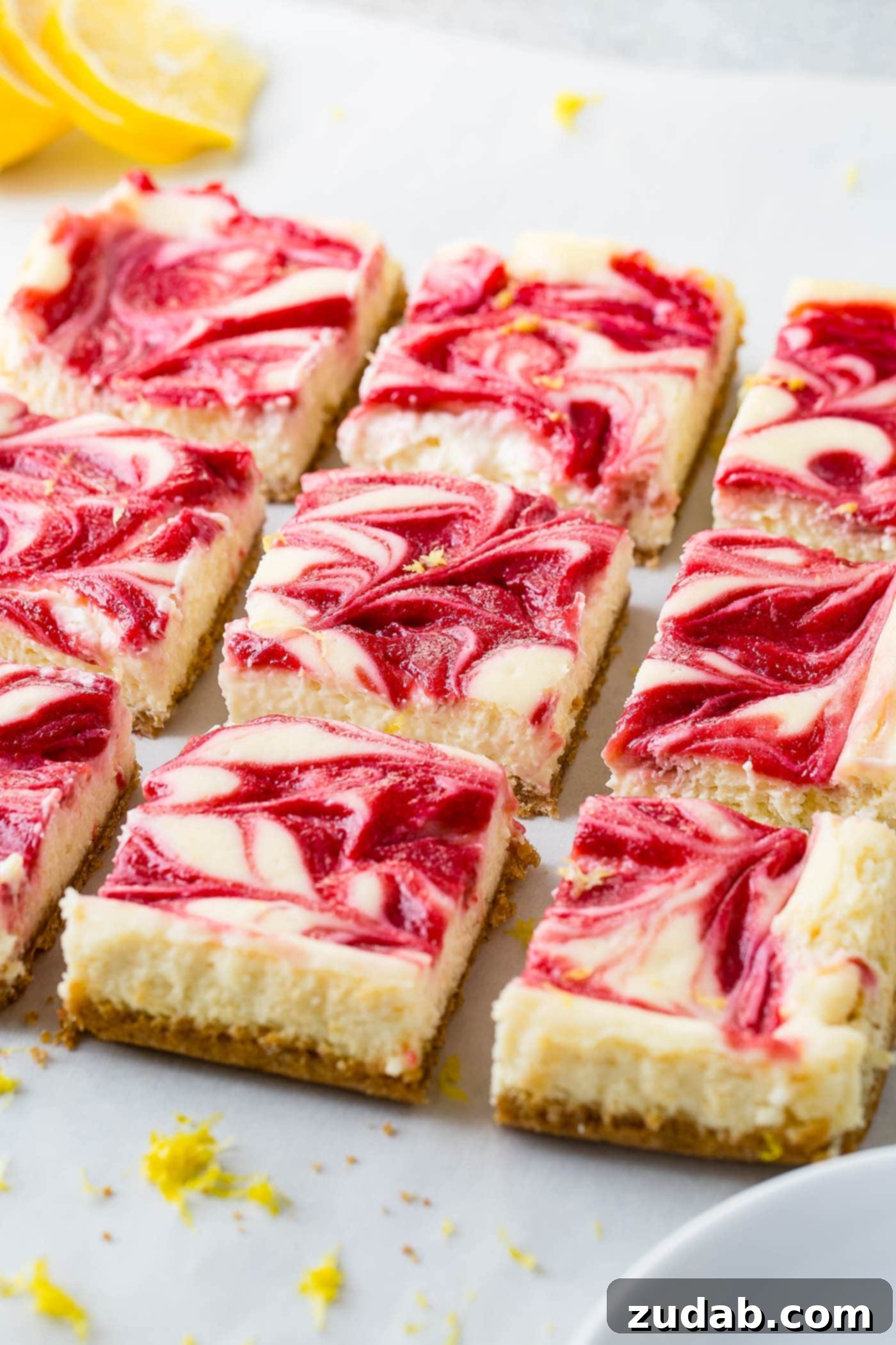 Zesty Berry Cheesecake Swirls 4 Nine precisely cut square portions of swirled raspberry lemon cheesecake bars. The visible layers include a golden graham cracker crust, a creamy, light yellow lemon cheesecake layer, and an elegant swirl of rich raspberry sauce adorning the top surface.