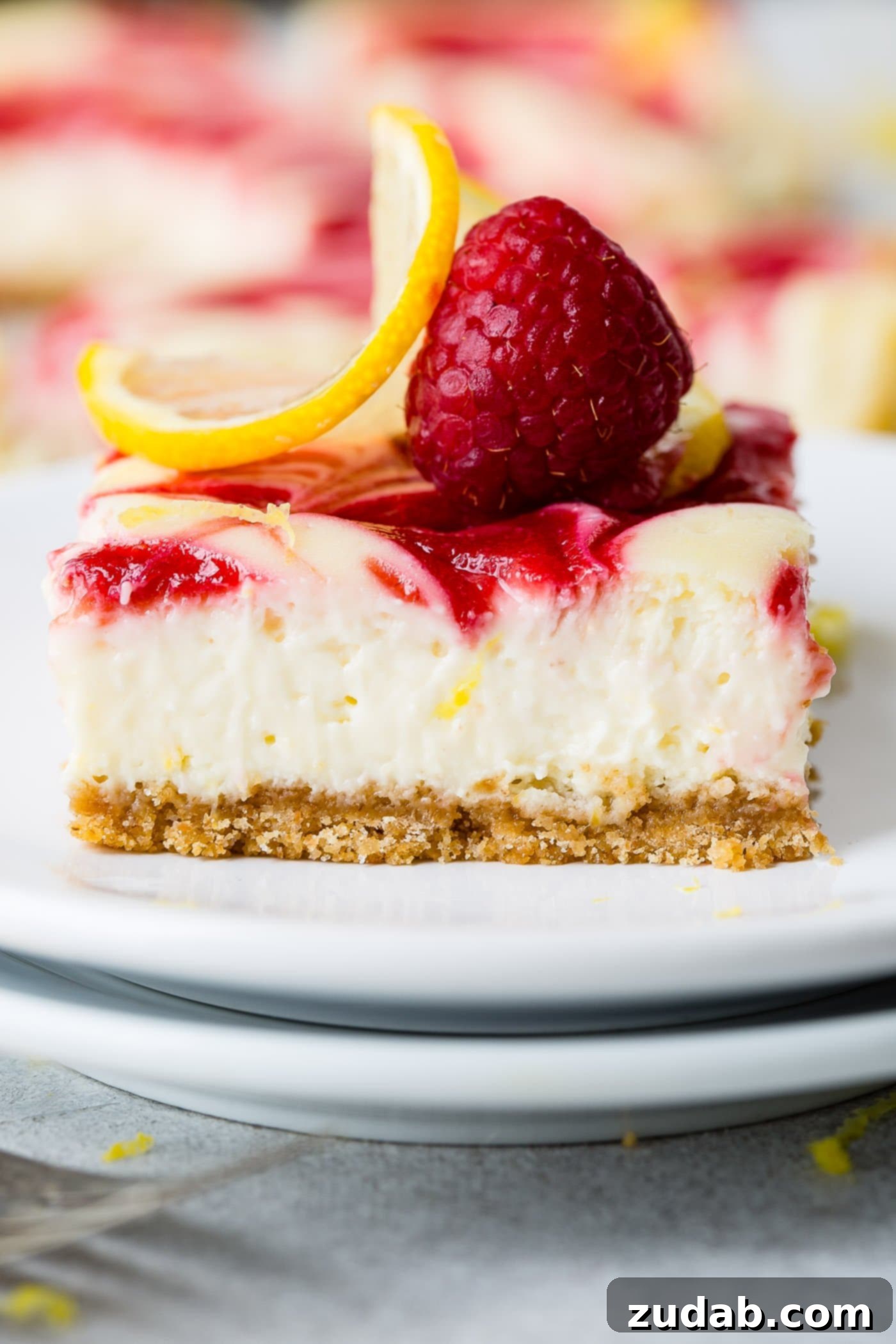 Zesty Berry Cheesecake Swirls 5 A perfectly portioned square of swirled raspberry lemon cheesecake rests on a white dessert plate. The golden graham cracker crust is visible, supporting the creamy lemon cheesecake filling topped with a striking raspberry swirl, garnished with a single whole raspberry and a delicate lemon slice.