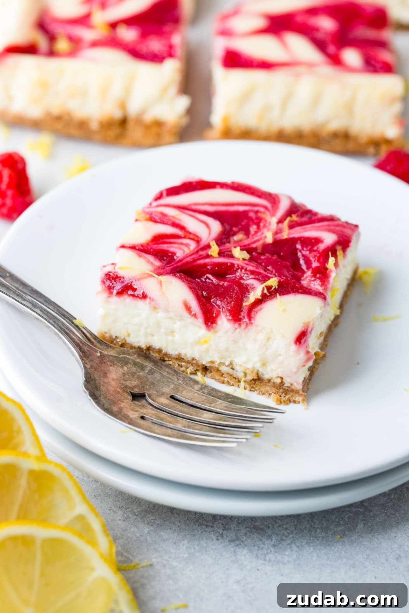 Zesty Berry Cheesecake Swirls 6 A single square of swirled raspberry lemon cheesecake presented on a white dessert plate, with a fork resting beside it. The cheesecake boasts a crisp graham cracker crust, a creamy lemon filling, and a beautiful, vibrant red swirl of homemade raspberry sauce on its top surface.
