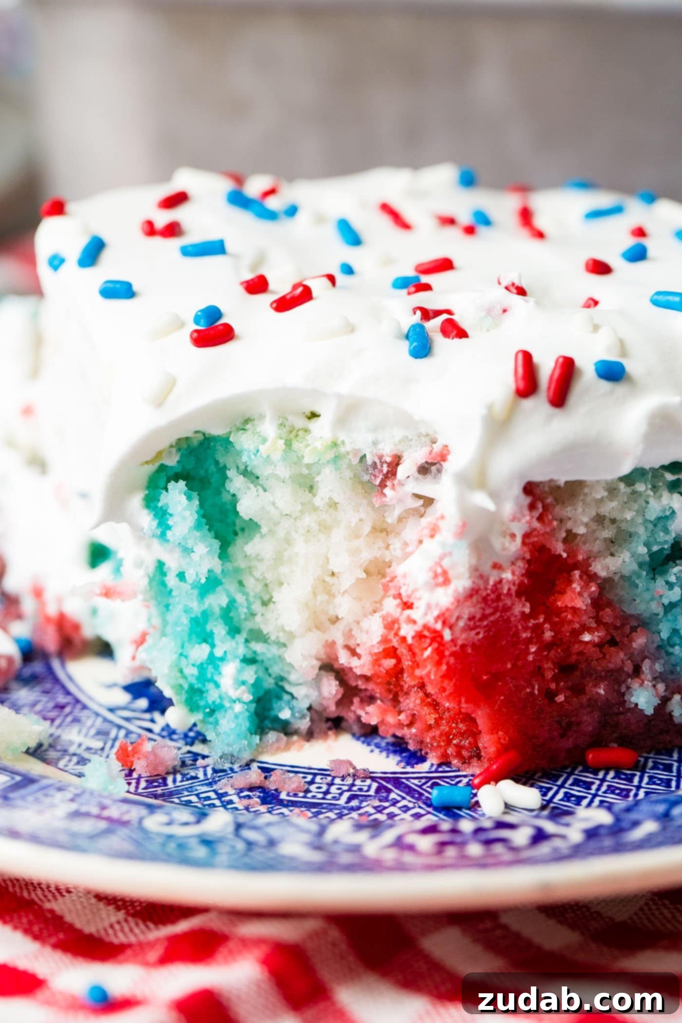 Patriotic Red White and Blue Jello Poke Cake 3 A piece of red, white, and blue poke cake. It is frosted with white frosting and sprinkled with red, white and blue sprinkles. The cake is white with areas of red and areas of blue.