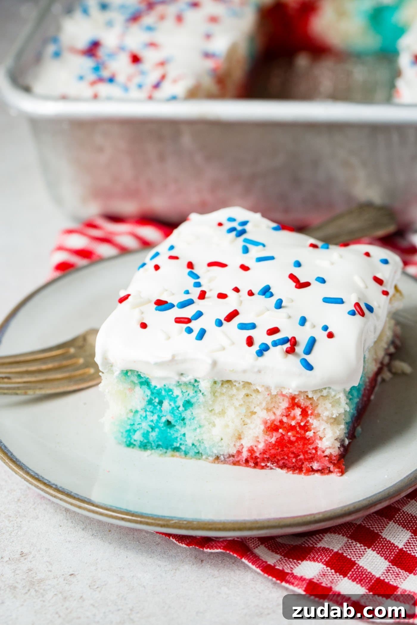 Patriotic Red White and Blue Jello Poke Cake 4 A piece of red, white and blue poke cake on a white dessert plate with a fork resting next to the cake. There is a red and white checkered napkin in the background.