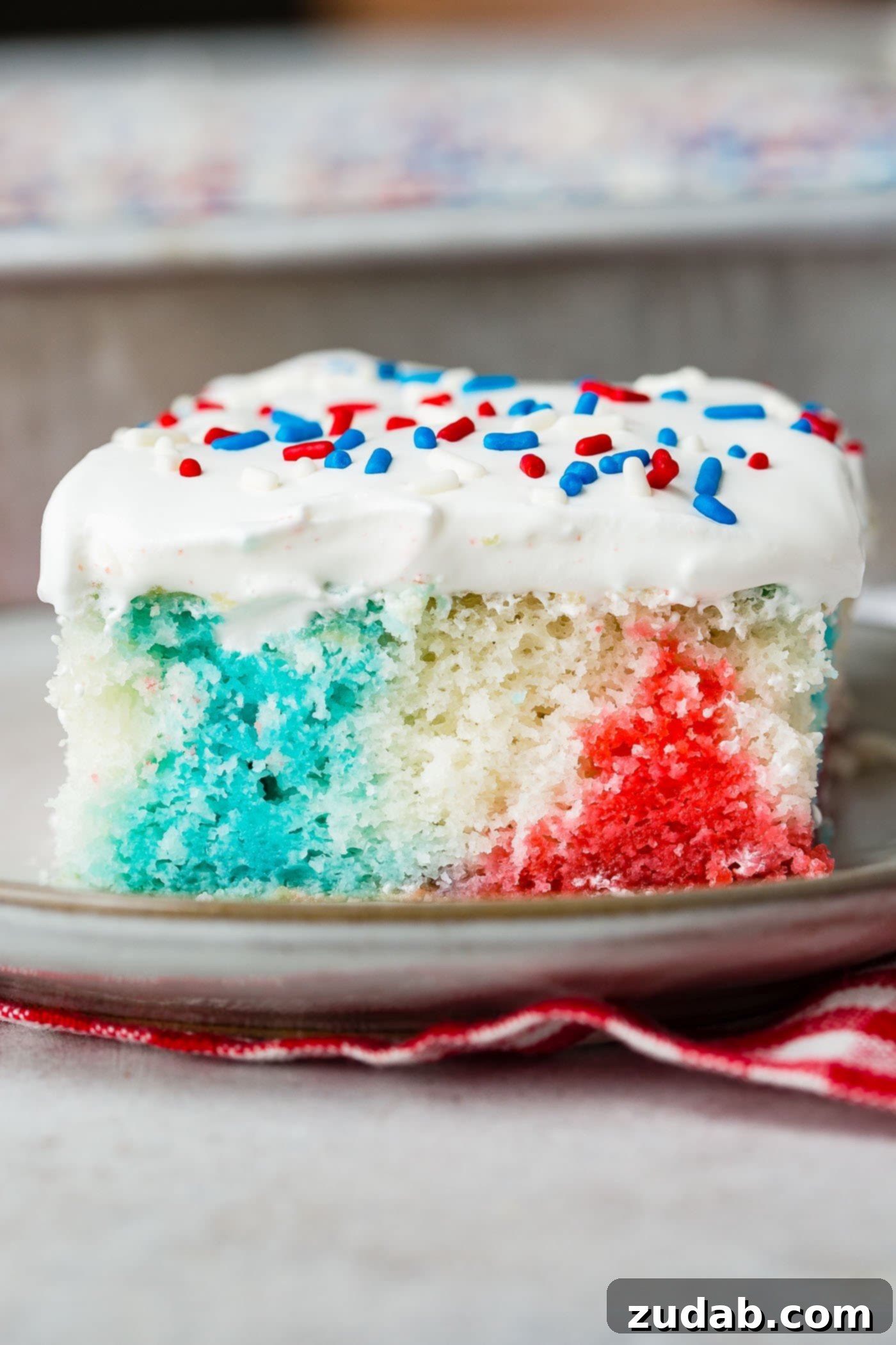Patriotic Red White and Blue Jello Poke Cake 5 A piece of red, white and blue poke cake on a dessert plate. The cake is frosted with white frosting and sprinkled with red, white and blue sprinkles.