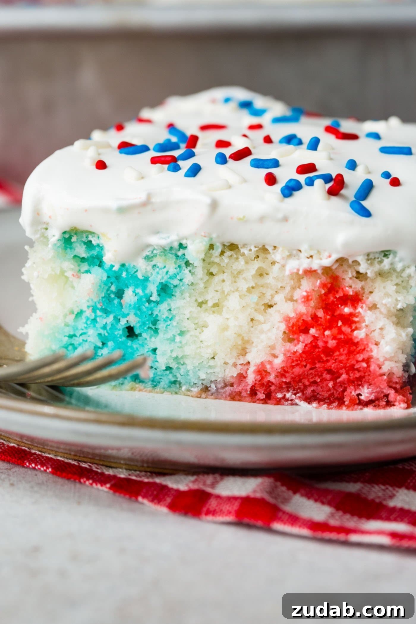 Patriotic Red White and Blue Jello Poke Cake 6 A piece of poke cake with a fork resting next to the cake. The cake is red, white and blue and is topped with white frosting and red, white and blue sprinkles. It is sitting on a red and white checked napkin.