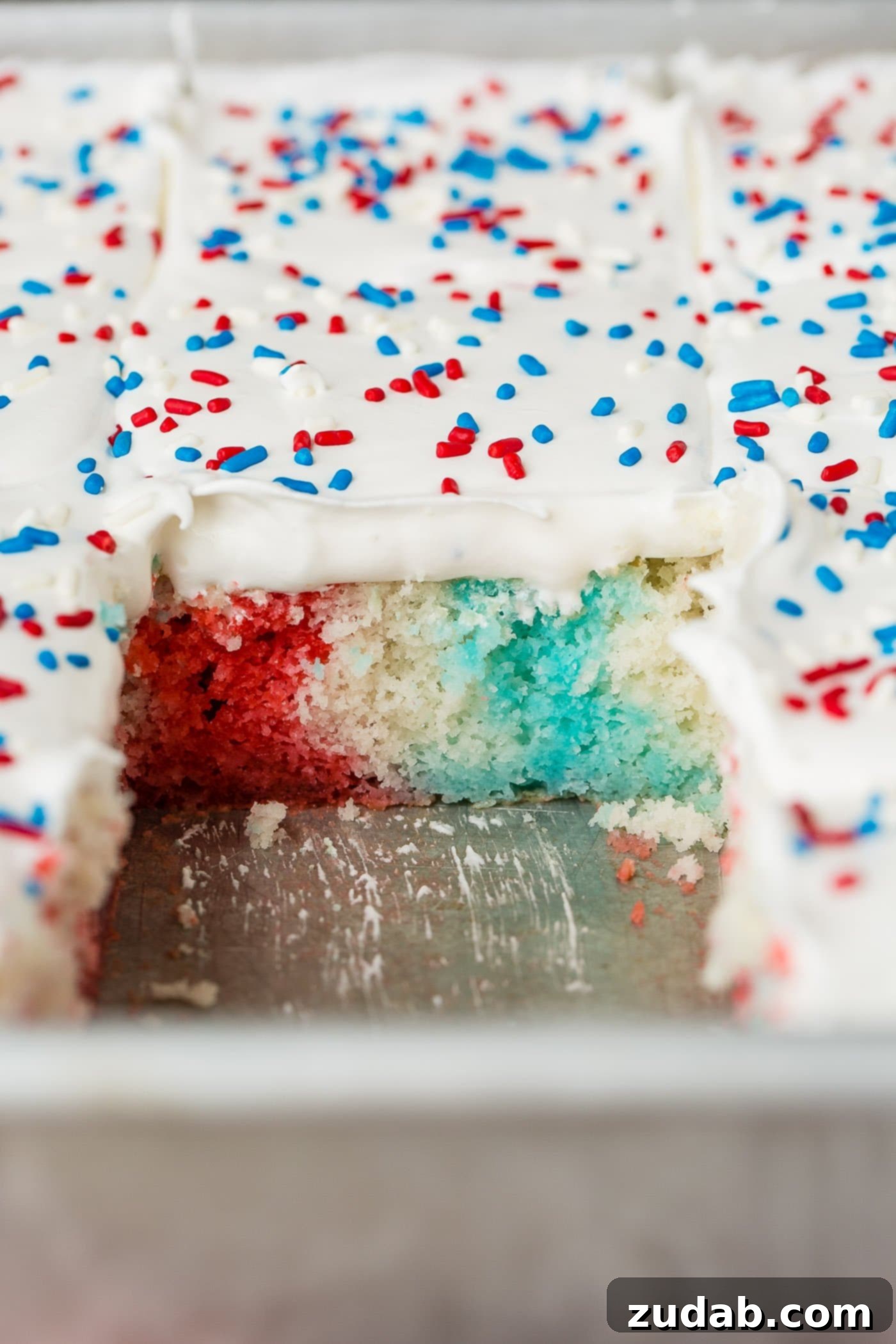 Patriotic Red White and Blue Jello Poke Cake 7 Red, white and blue cake with a slice removed exposing the red, white and blue inside of the cake. The cake has white frosting and red, white and blue sprinkles on top.