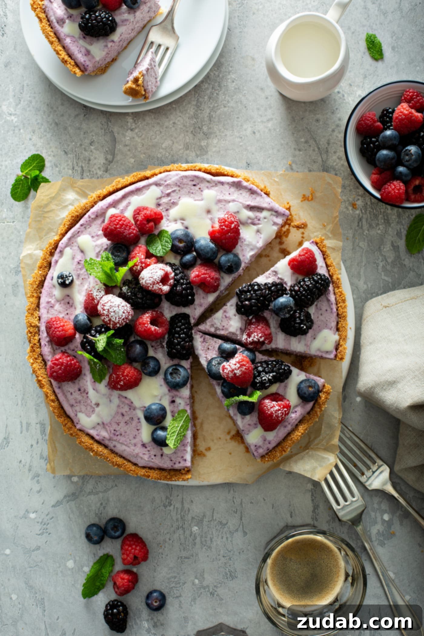 White Chocolate Berry Icebox Pie 2 A berry white chocolate pie that has been sliced. A slice has been removed and two more slices are separated. The pie has a graham cracker crust and is topped with strawberries, blue berries and raspberries. A few mint leaves are scattered on top of the pie and on the table next to the pie. There are two forks, a napkin and a bowl of mixed berries on the table.