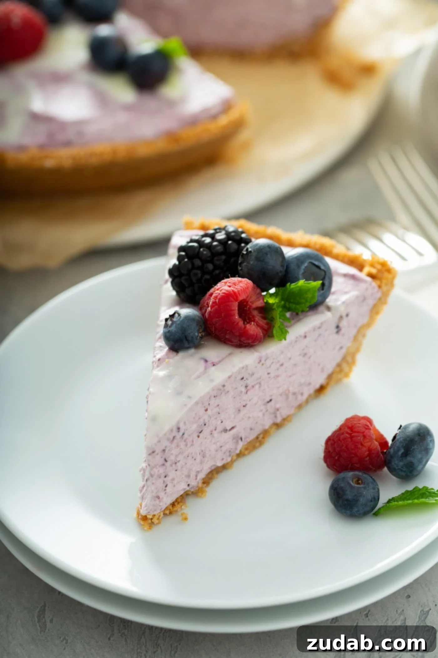 White Chocolate Berry Icebox Pie 11 A white dessert plate with a slice of white chocolate berry pie. The pie is pink in color and you can see bits of pureed berries in the filling. the pie has raspberries, blue berries and black berries and a mint leaf on top. There are berries on the plate and a white chocolate berry pie and a couple of forks in the background