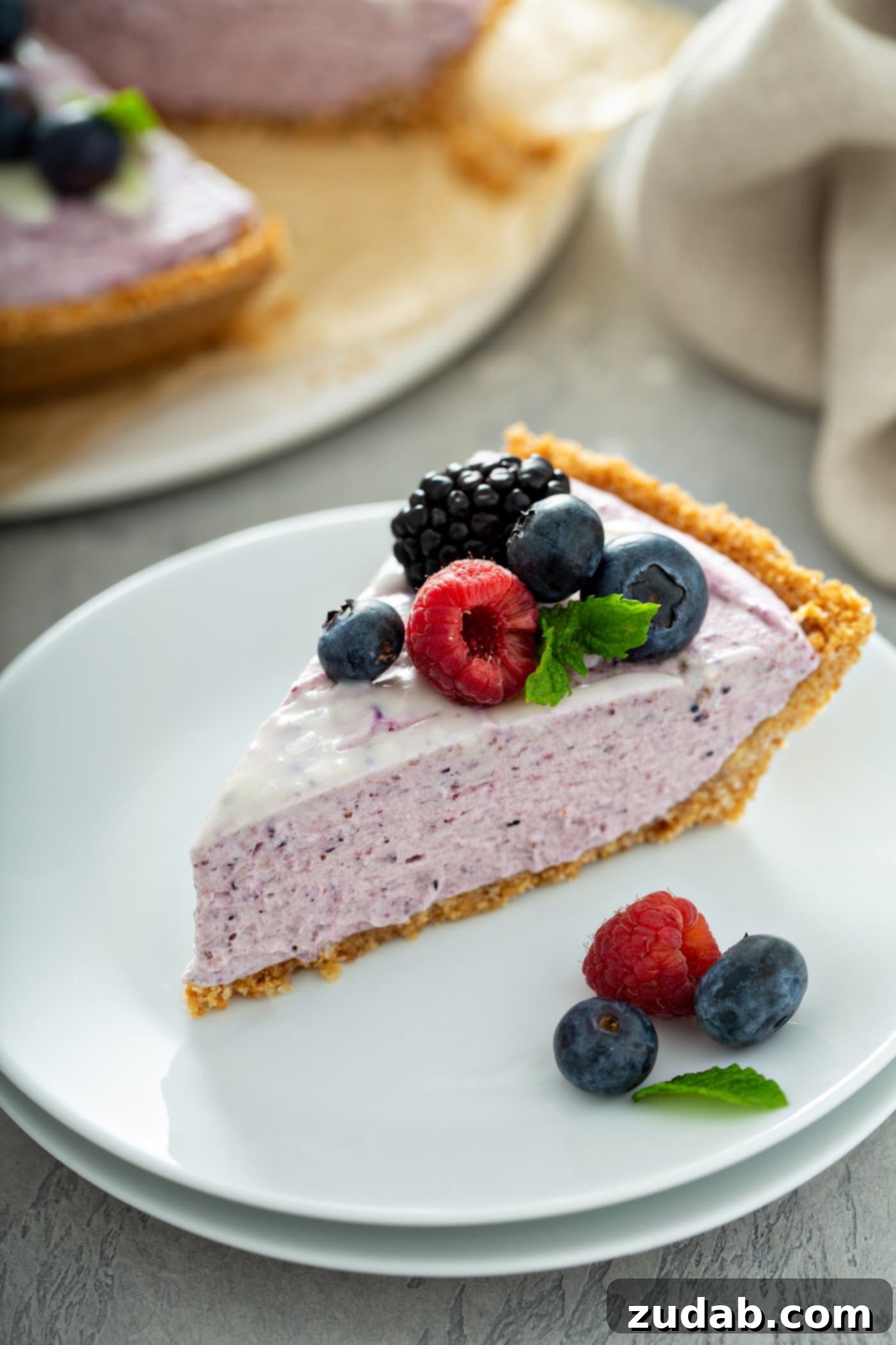 White Chocolate Berry Icebox Pie 14 A slice of white chocolate berry pie on a dessert plate. There are a few mixed berries and mint leaves on the plate and the pie slice is topped with raspberries, blue berries and black berries. A mint leaf is next to the berries and a white chocolate sauce is drizzled on top of the pie. A pie plate with a whole pie and a napkin are in the background.