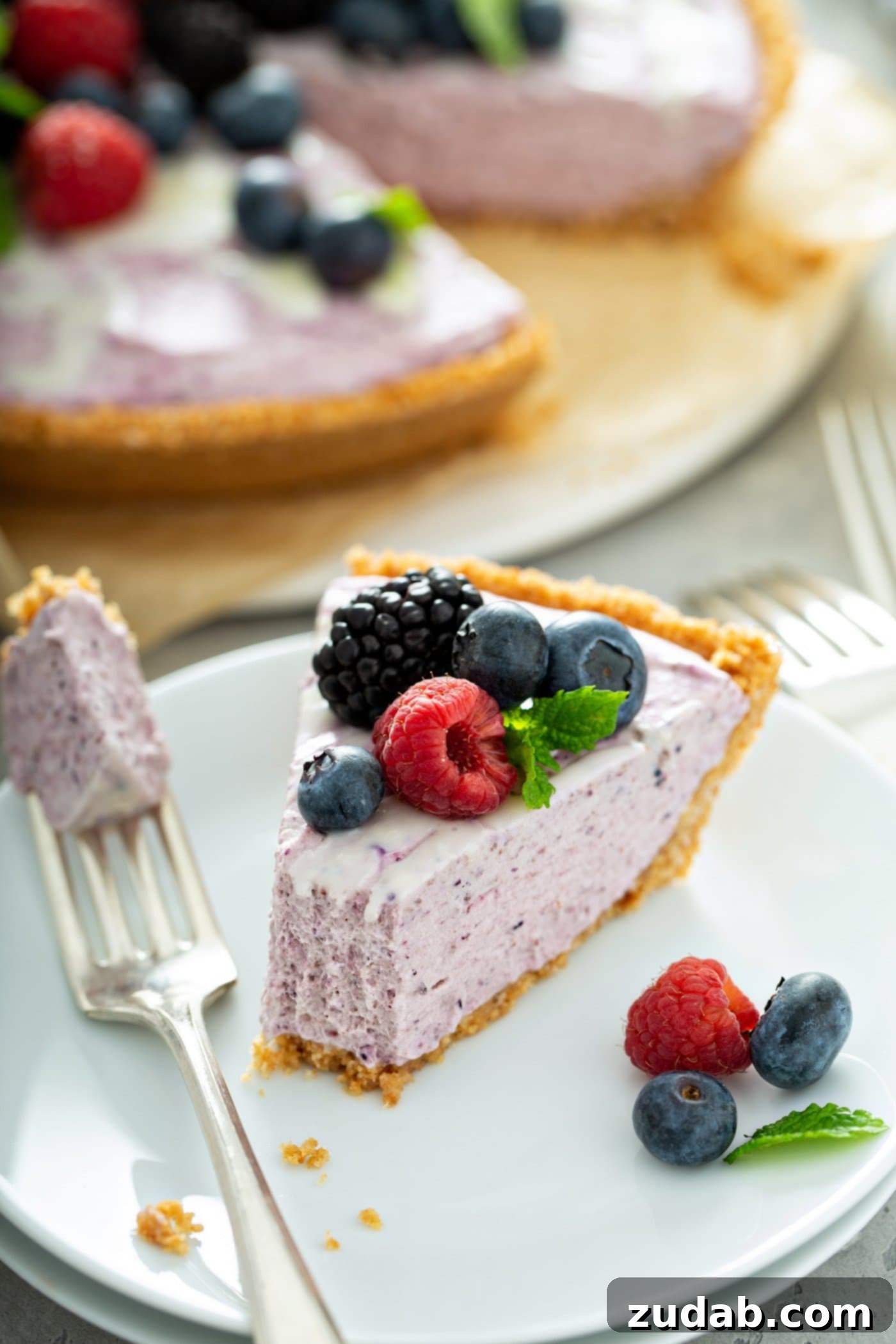 White Chocolate Berry Icebox Pie 3 A slice of white chocolate berry pie on a dessert plate. A fork with a bite-size piece of pie on it is next to the slice of pie. the pie is topped with raspberries, black berries and blue berries and one mint leaf. there are berries on the plate next to the pie and the whole pie in which a slice a has been removed, is in the background.